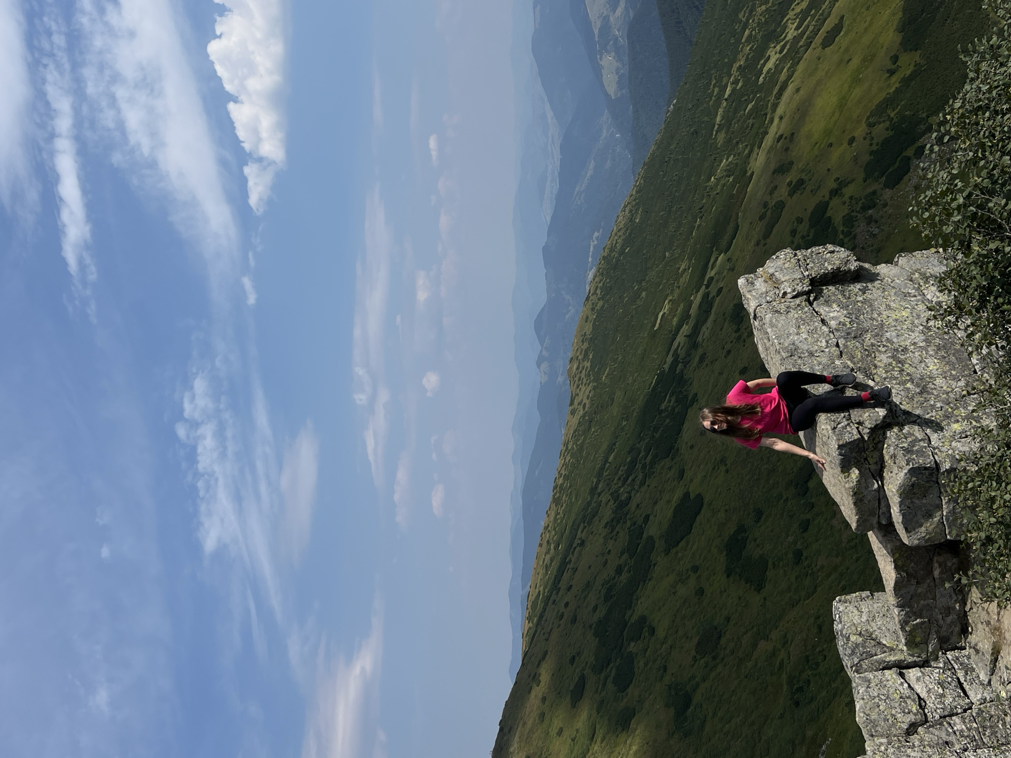 A young woman with blonde hair, wearing a pink t-shirt and black leggings, sits on a rocky outcrop overlooking a vast mountain landscape. The mountains are covered in green vegetation and stretch into the distance, appearing in shades of blue and green due to atmospheric perspective. The sky is a clear blue with scattered white clouds. The woman is positioned on the right side of the frame, with her legs dangling over the edge of the rock. The rocky formation is large and textured, with visible cracks and moss. This scene is located in the Carpathian Mountains near Vorokhta, Ukraine.