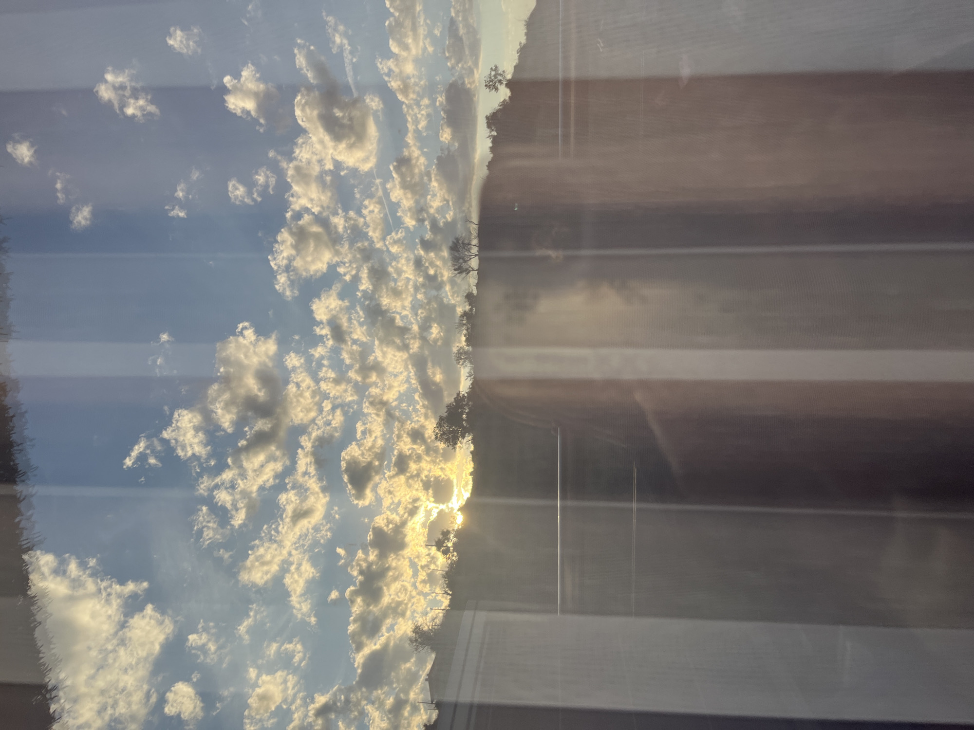 The image captures a scenic view of a sunset over a hilly landscape, seen through a window with vertical blinds. The sky is filled with scattered clouds, some illuminated by the golden rays of the setting sun, which is partially obscured by the terrain. The sun's light creates a vibrant glow and casts rays across the sky. The hills in the background are covered with vegetation, and a few trees are visible against the horizon. In the foreground, the window frame and vertical blinds are evident, creating a striped effect that overlays the outdoor scene. There are reflections on the window, including what appears to be a faint outline of a person. The overall atmosphere is peaceful and serene, indicative of an evening sunset. The location is specified as Varna, Bulgaria.