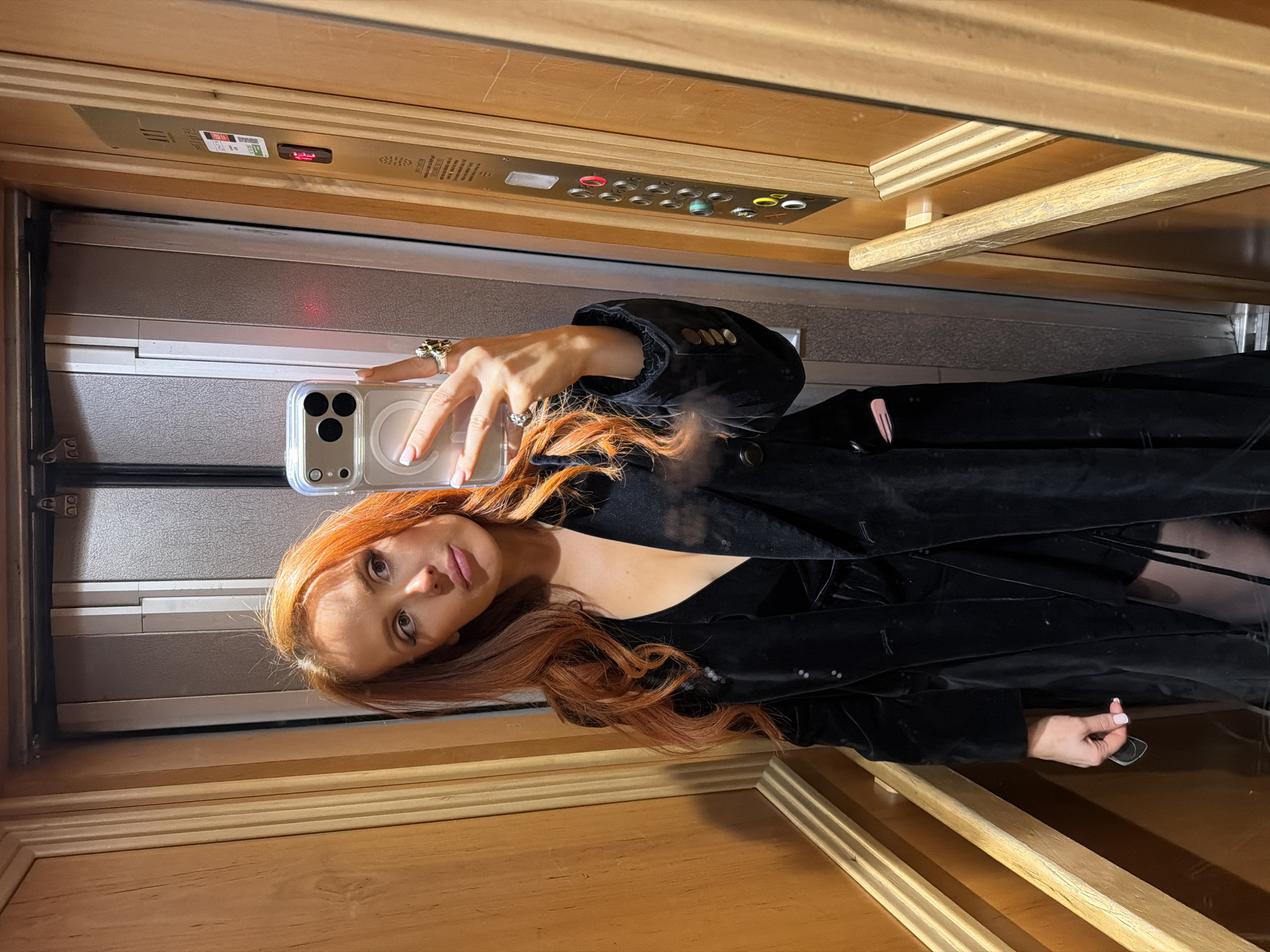 A female individual with long, wavy auburn hair is observed inside an elevator, taking a selfie using a smartphone held in her right hand. She is wearing a dark, possibly velvet or sequined, long-sleeved blazer/coat over a dark, low-cut garment. Dark hosiery is visible on her legs. Her right hand displays multiple rings and light-colored manicured nails. Her left hand holds a small, dark object, possibly a key card.
The elevator interior features light-toned wooden paneling with vertical grooves and horizontal wooden handrails. The elevator doors, visible in the reflection, appear metallic. A control panel on the right wall contains a digital display showing the number '1' in red, along with several circular buttons, some illuminated. A sticker below the display shows a QR code and partially visible French text, "VERIFICATION PERIODIQUE..." The letters "ATT" are visible at the top of the panel.
The presence of French text on the elevator control panel indicates the location is in France. As requested, the scene is located within an elevator in the city of Paris, France. The exact building or street address is not discernible from the image.