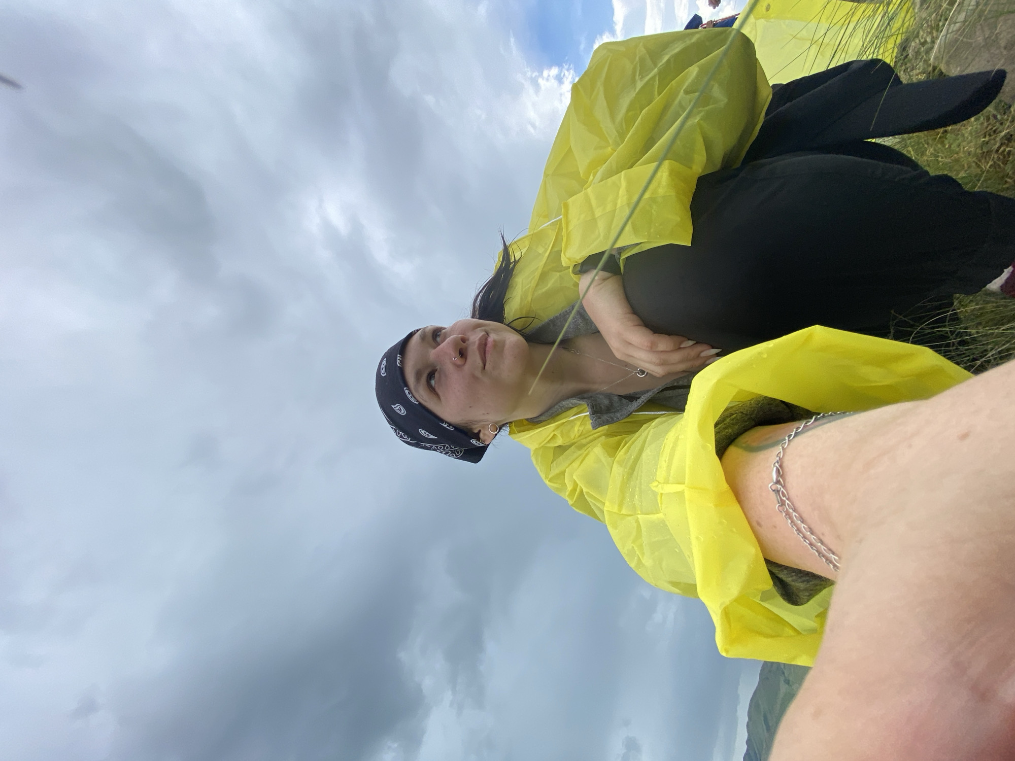 A low-angle, medium shot captures a young person from the chest down, wearing a bright yellow, crinkled raincoat and black pants. They are crouched down on a grassy hillside under a dramatic, cloudy sky. The person wears a black bandana with a white paisley pattern on their head, a nose ring, and a silver necklace with a circular pendant. A silver chain bracelet is visible on their left arm. Their gaze is directed upwards and to the right, towards the sky. The background shows rolling, green hills and a sliver of blue sky peeking through the clouds. The image appears to have been taken outdoors in a rural or natural setting, possibly during overcast or rainy weather. The specific location in the city of Рыскулово, Russia, is not discernible from the visual information provided in the image.