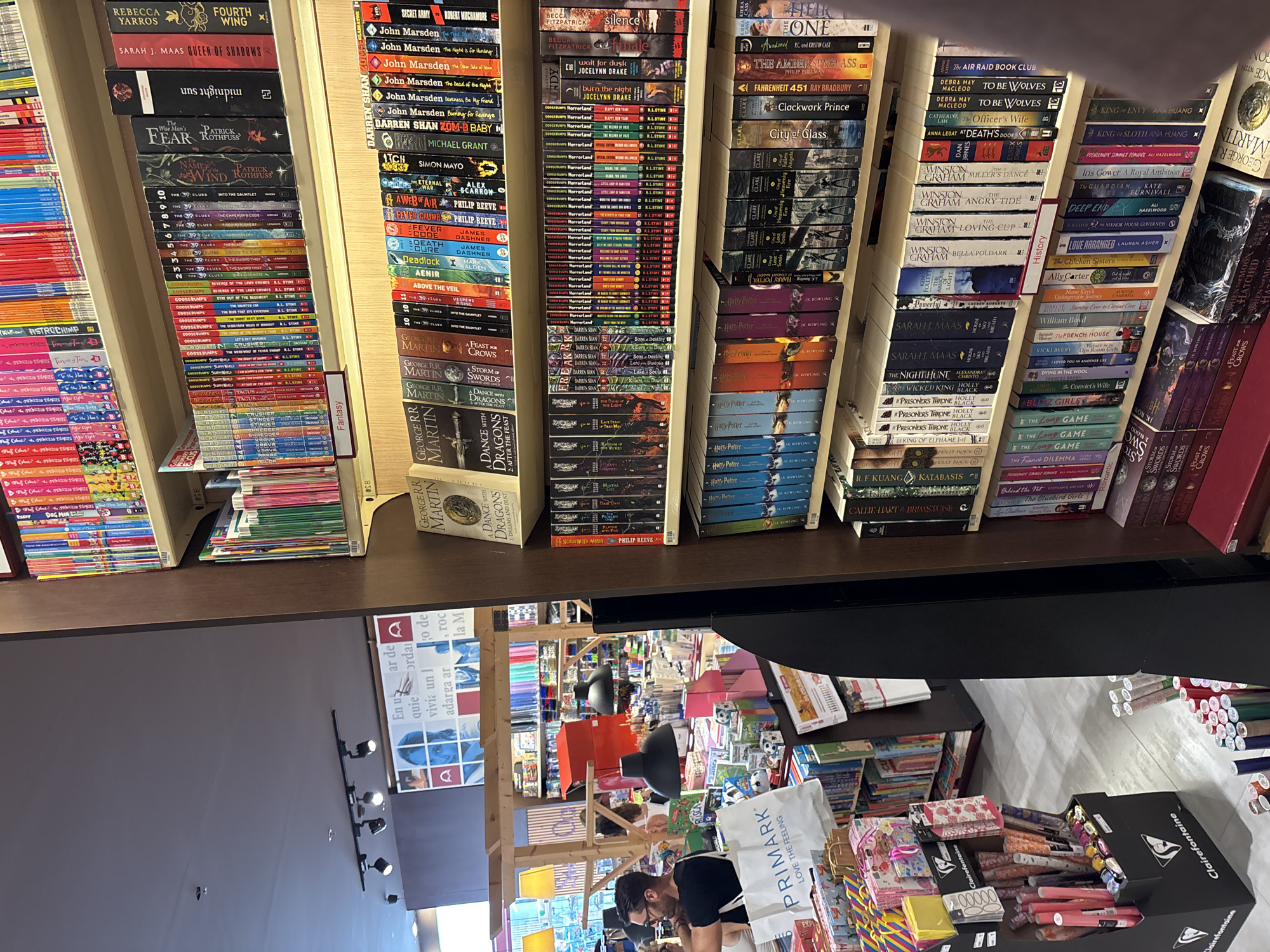 The image depicts the interior of a bookstore in Orihuela, Spain. A large section of a wooden bookshelf is filled with various books. On the top shelf, a row of colorful children's books is visible, followed by a section of fantasy novels with titles like "FEAR" and "QUEEN OF SHADOWS." Below that, more fantasy and young adult books are displayed, including a large collection of "The 39 Clues" series. The bottom shelf showcases more books, including "Harry Potter" series, and various other novels. In the background, a blurred image shows a store interior with people browsing and displays of stationery and gift items, including "Primark" branded bags. The lighting appears to be artificial, coming from overhead fixtures.