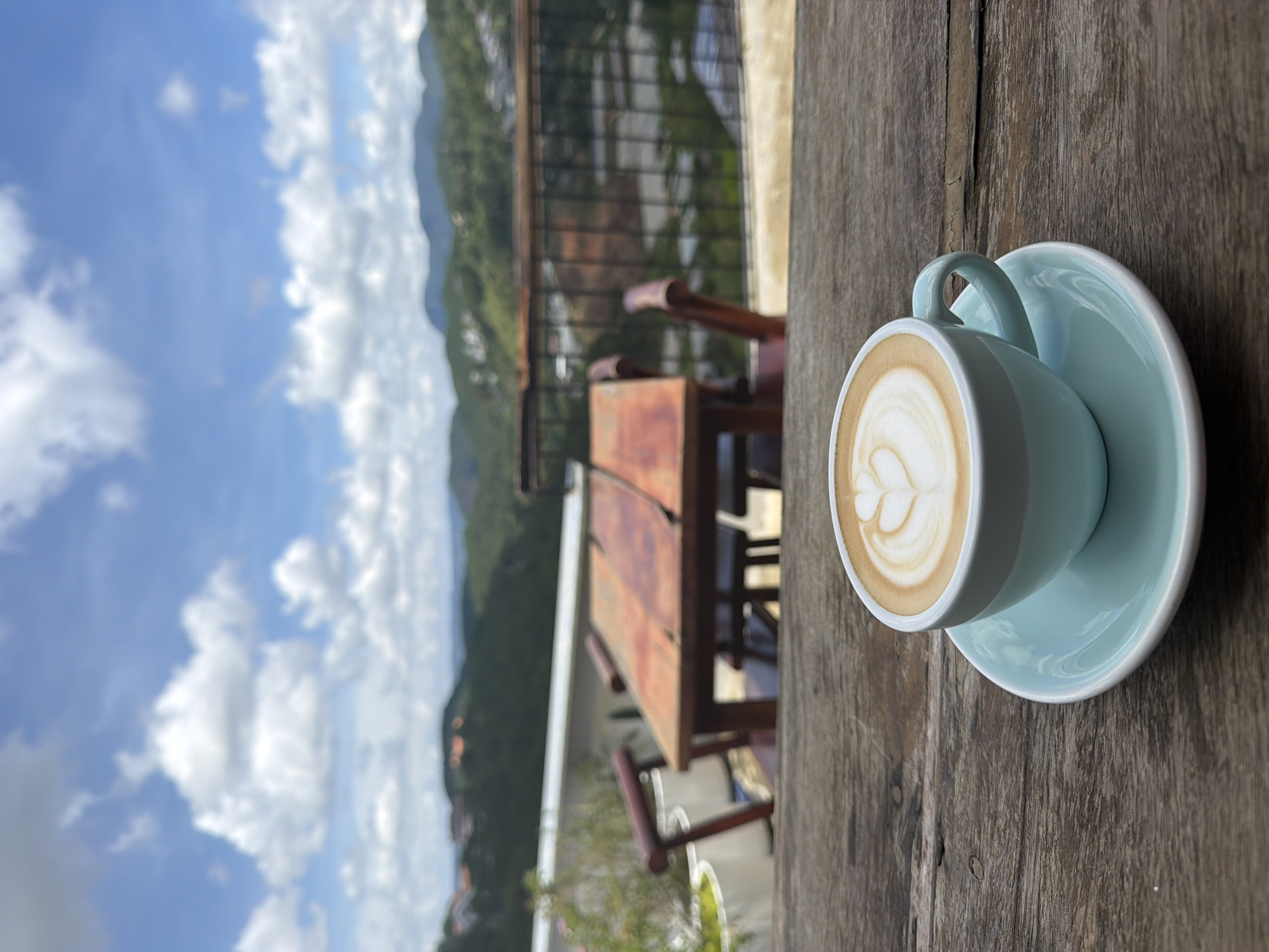 A close-up, eye-level shot captures a light blue ceramic cup filled with cappuccino on a matching saucer, positioned on a weathered wooden table. The coffee features latte art in the form of a heart shape. In the background, out of focus, are outdoor seating arrangements including a wooden table and chairs, a railing, and a lush green mountainous landscape under a bright blue sky with scattered white clouds. The scene is likely at a cafe or restaurant in Da Lat, Vietnam, known for its scenic mountainous terrain and cool climate, offering a picturesque view to patrons.