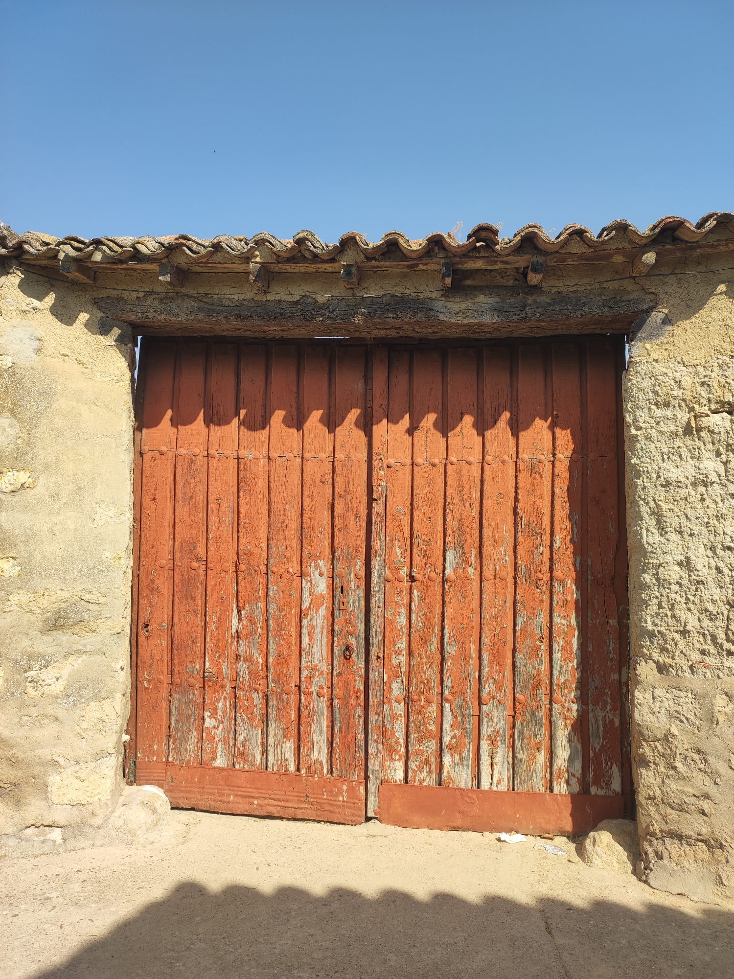 A rustic, weathered, double wooden door, painted a faded orange, is the central focus of the image. The door is set within a stone and stucco archway, topped by a terracotta tiled roof. The wood of the door shows significant wear, with peeling paint revealing the natural grain beneath. Metal studs are visible along horizontal bands across the door. A dark shadow is cast on the ground in front of the door, indicating strong sunlight. The environment appears to be outdoors, possibly in a rural or historic setting, with a clear blue sky overhead. A small, dark object, possibly a bird, is visible in the sky above the roof. The overall impression is one of age and simplicity.