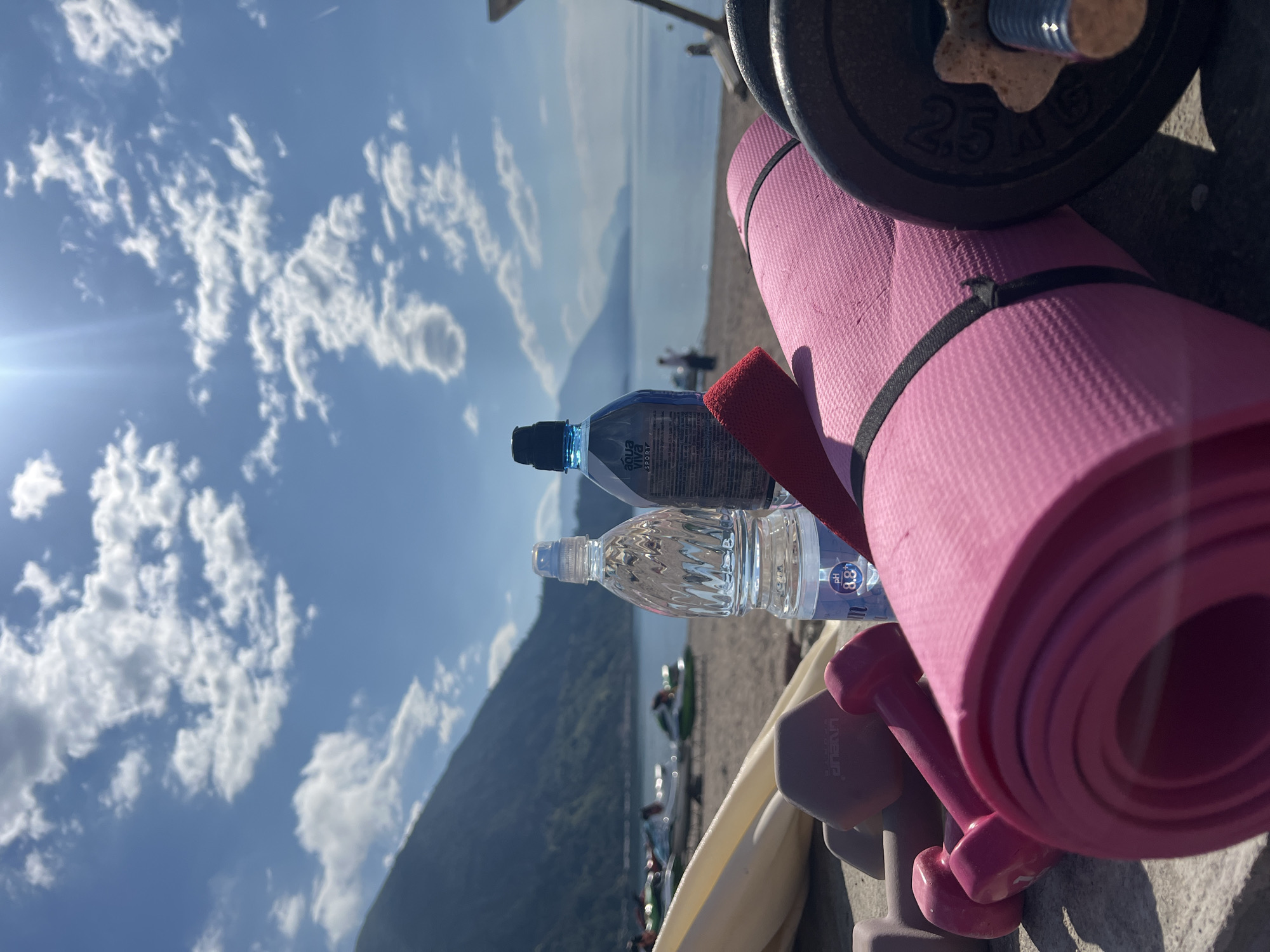 The image shows a scene at the beach in Prijevor, Montenegro. Exercise equipment (pink yoga mat, dumbbells, weight plate) and two water bottles are arranged on a concrete surface. In the background, the beach stretches to the edge of a lake surrounded by mountains. The sky is bright blue with puffy clouds. Several people are seen in the middle distance near the water.