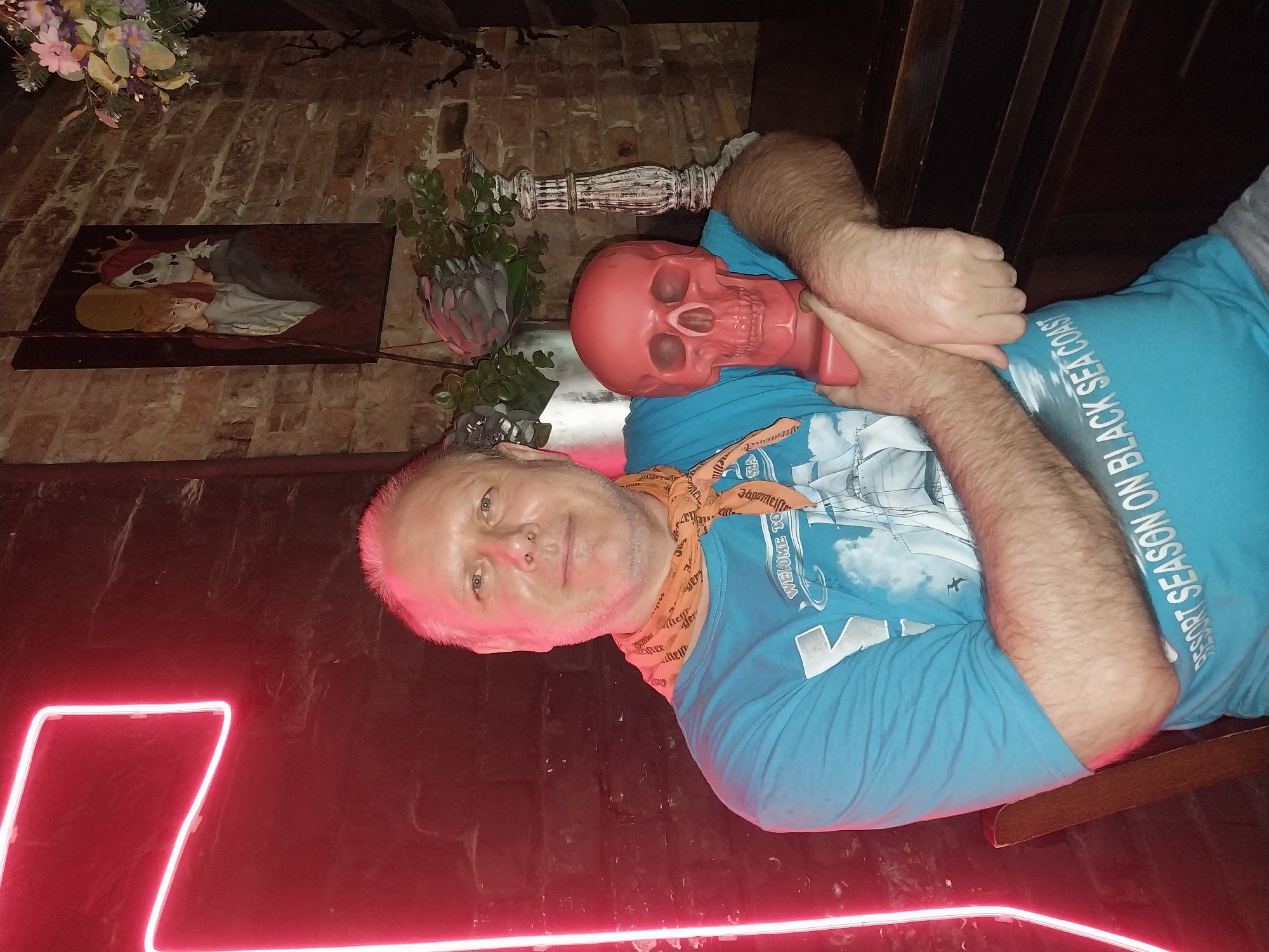 The image shows a man in a blue t-shirt with an orange bandana around his neck. He is holding a red skull in his arms. He is sitting in a dimly lit room with brick walls. There is a neon light sign in the background that appears to be a large "T". A painting of two figures, one human and one skeletal, hangs on the wall behind the man. There are also some plants and a candlestick visible. The man is smiling and looking at the camera. The overall atmosphere of the image is dark and slightly spooky. It is impossible to determine the location in the city of Orenburg, Russia based on the provided image.