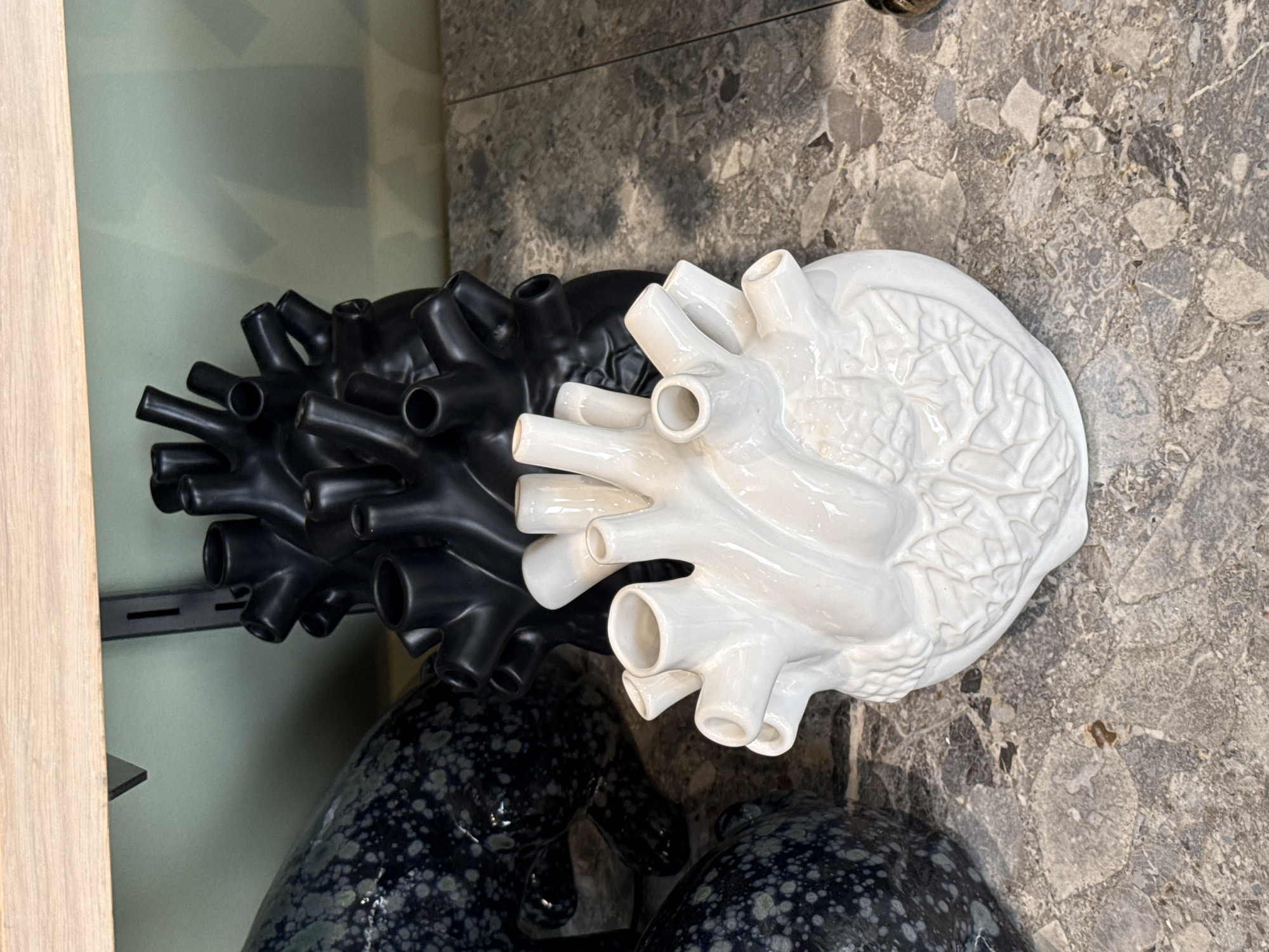 The image shows two decorative heart-shaped objects, one white and one black, placed on a gray marble or stone surface. The objects are detailed and appear to be made of ceramic or a similar material. They are anatomical representations of a human heart, with multiple tubes protruding from the top. The black heart is positioned slightly behind and to the left of the white heart. A section of what looks like a wooden shelf or wall is visible on the left edge of the frame. The background suggests an indoor setting, possibly a store or showroom. There is no evidence to suggest the location is in Cape Town, South Africa.