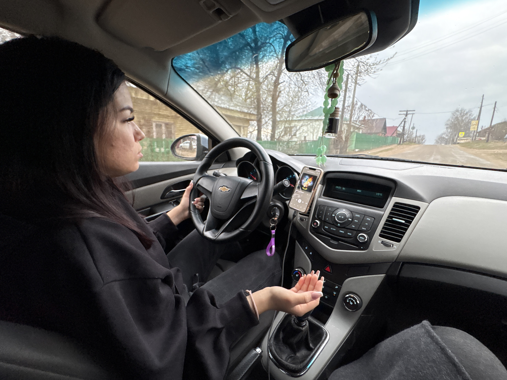 A woman with dark hair is driving a Chevrolet vehicle in the town of Фоки, Russia. She is holding the steering wheel with her left hand, and her right hand is near the gear stick. The vehicle's dashboard is visible, including the radio and phone holder. Outside the windshield, there is a street with houses and utility poles. The sky is overcast.