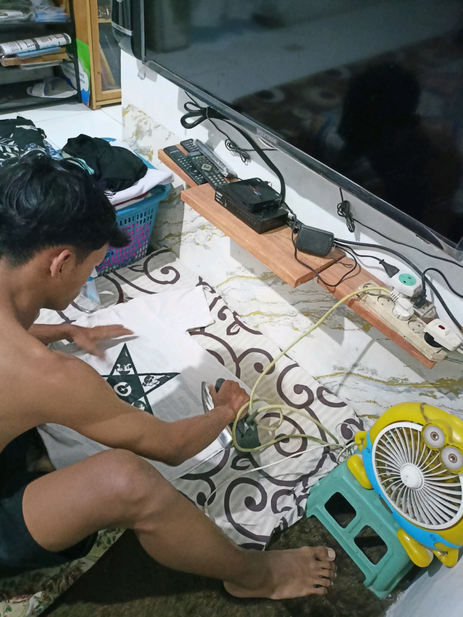 In Payaman, Indonesia, a young man is seen ironing a white t-shirt with a black graphic design on it. The t-shirt is laid out on a patterned bedspread. The man is positioned on his knees, with his back partially to the camera as he applies the iron. Next to him, a small yellow fan shaped like a Minion character sits on a teal plastic stool. In the background, a television screen is visible, along with a wooden shelf holding electronic devices and remote controls, and a blue laundry basket filled with clothes. The environment suggests an indoor setting, possibly a bedroom or living area.