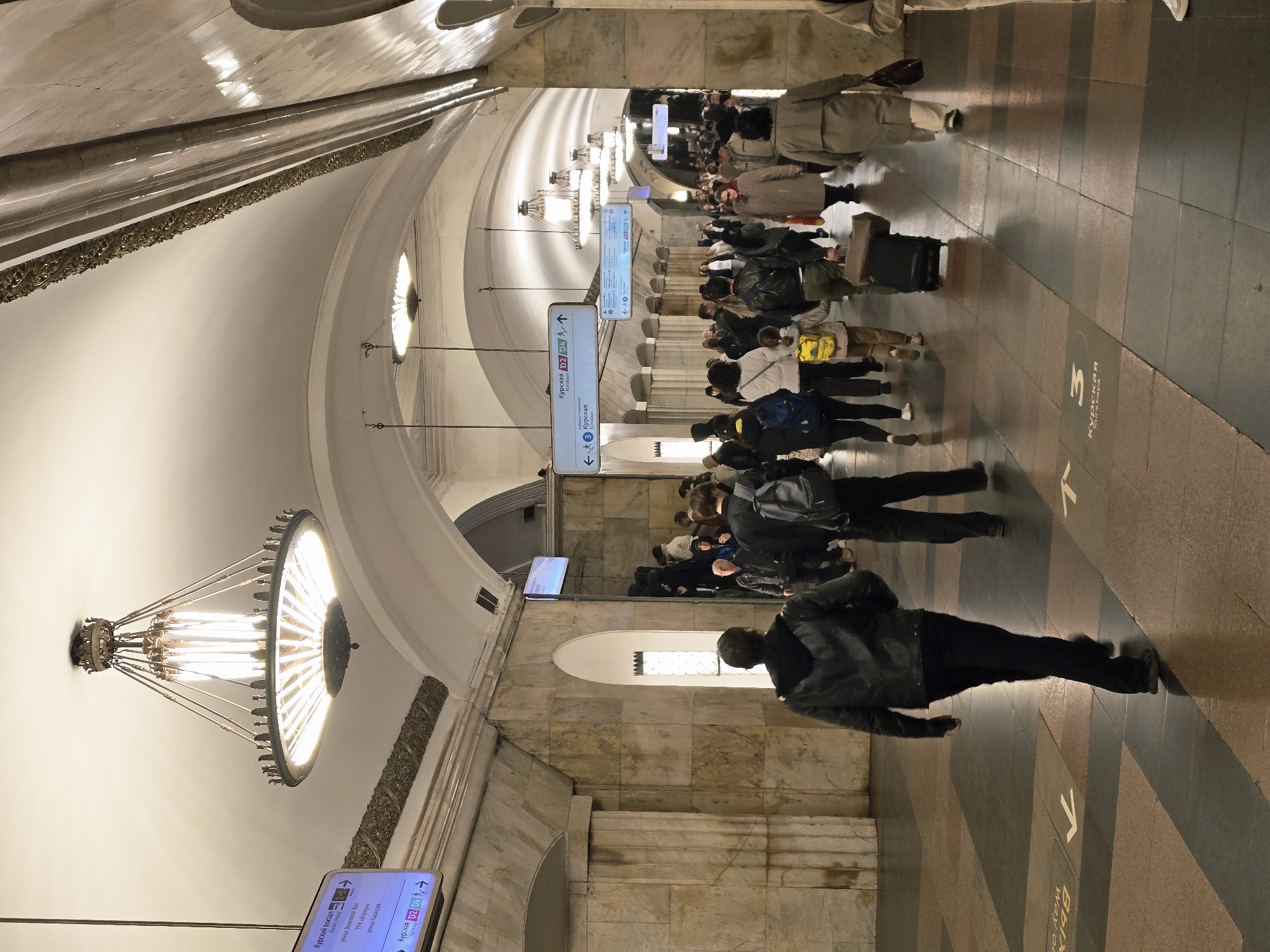 The image shows an indoor scene, likely a subway station in Moscow, Russia. A group of people is walking along a wide platform, their movements blurred suggesting they are in motion. Signage in Russian is visible, with one sign indicating the direction to Kurskaya station and another mentioning Kurskiy Vokzal (Kursky Station) and local streets. The station's architecture is characterized by high ceilings, ornate lighting fixtures, and marble walls. The floor is patterned with stripes and arrows. A man is walking in the foreground.