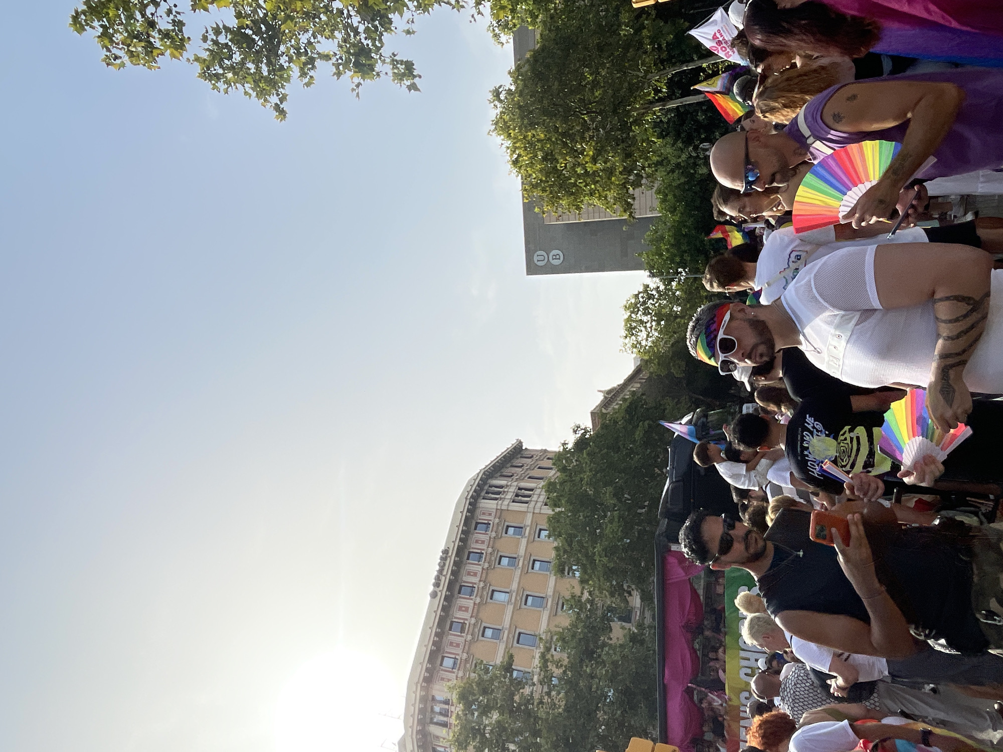 This image captures a street scene in Barcelona, Spain, during what appears to be a pride event. The foreground features several individuals, including a man in a black sleeveless shirt and sunglasses engrossed in his phone, and another man wearing a white mesh shirt, sunglasses, and a rainbow headband. This second man holds a colorful fan, an item also held by a bald man in a purple tank top and sunglasses to his right. In the background, a crowd of people are visible, some with rainbow flags, suggesting a celebratory atmosphere. Buildings characteristic of Barcelona's architecture are present, with one being a light-colored, multi-story building with many windows, and another a darker, modern structure with a "U B" logo. Trees line the street, and the sky is bright blue, indicating daytime.