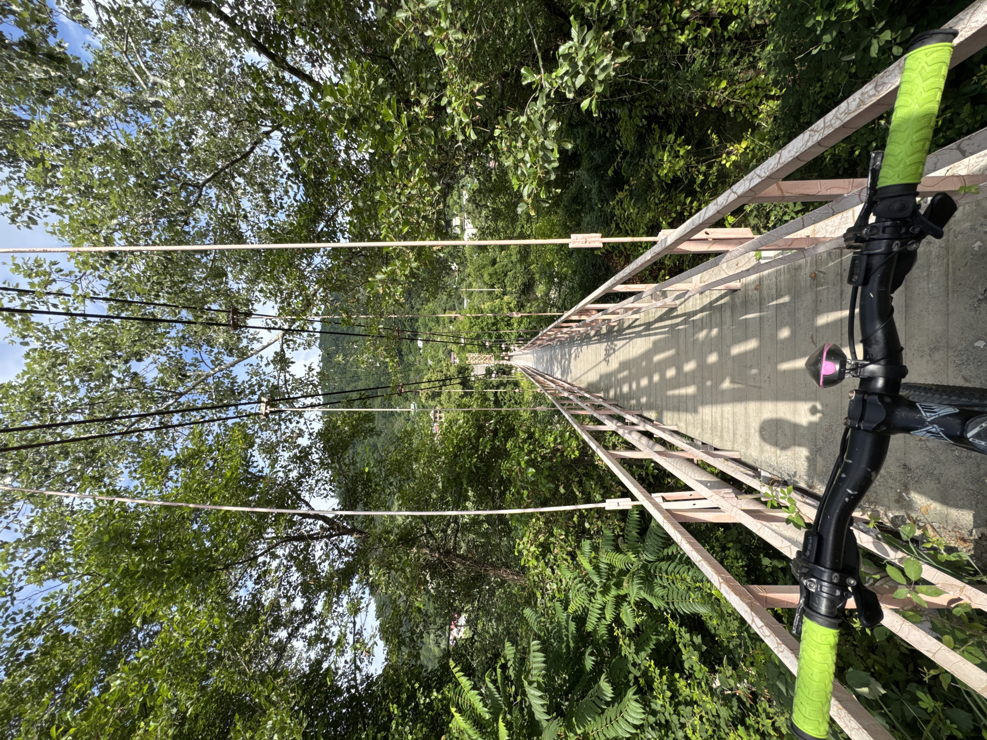 An eye-level, point-of-view shot captures a suspension bridge crossing a wooded area. The bridge, constructed of light-colored wood planks with pink railings, stretches into the distance, disappearing behind trees. The handlebars of a bicycle with bright green grips are visible in the foreground, suggesting the perspective of a cyclist. Lush green foliage crowds both sides of the bridge, with tall trees reaching towards a bright blue sky dotted with clouds. The scene is set in Sochi, Russia, likely in a natural park or along a scenic route, given the dense vegetation and the presence of a pedestrian and cyclist-friendly bridge. The lighting indicates a sunny day, casting distinct shadows on the bridge planks.