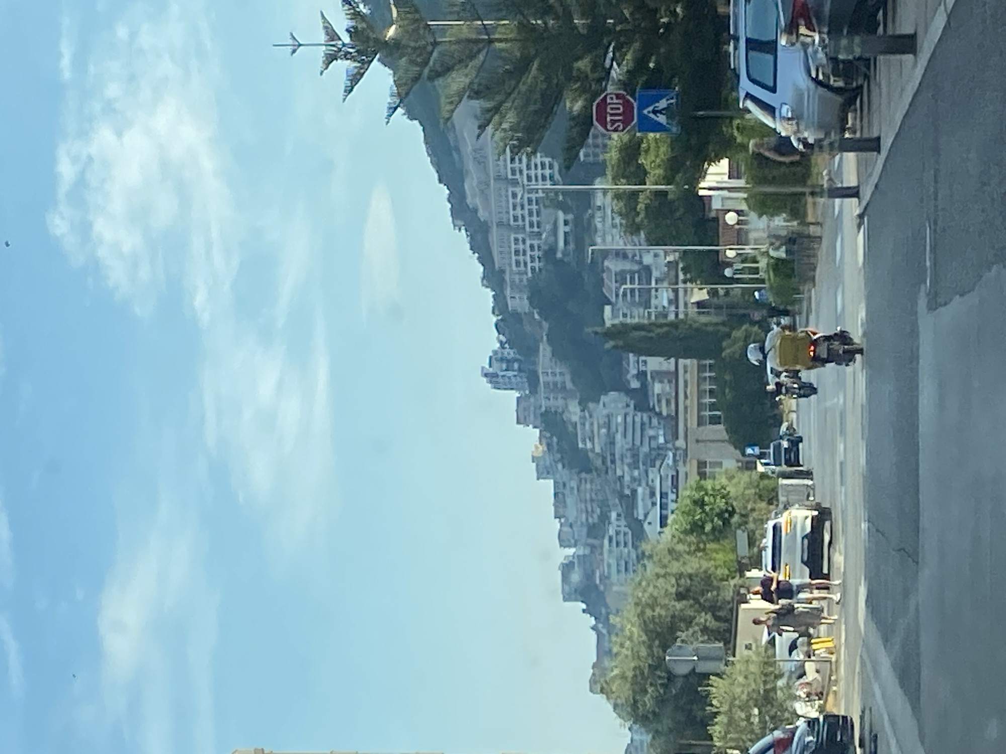 The image depicts a street scene in Budva, Montenegro, characterized by a steep hillside densely populated with buildings and lush greenery. The day appears to be bright and sunny, with a clear blue sky dotted with wispy clouds, suggesting fair weather.
On the right side of the frame, a road runs parallel to the hillside. Several vehicles are present, including a silver SUV parked near a lamppost and a white van further down the road. A motorcycle with a rider wearing a yellow jacket and helmet is visible, heading away from the viewer. Other people can be seen on the sidewalk, some standing and interacting, others seemingly waiting.
A prominent octagonal red stop sign is displayed on a pole, accompanied by a blue directional sign with a white symbol, indicating traffic regulations. The architecture of the buildings cascading down the hill suggests a Mediterranean or Balkan influence, with many structures featuring white facades and stacked levels. The dense vegetation of trees and bushes adds to the vibrant, natural setting.
The overall impression is of a bustling urban environment integrated into a dramatic natural landscape. The image captures a moment in time on a sunny day, with everyday activities of traffic and pedestrian movement unfolding against the backdrop of the striking Montenegrin coast.