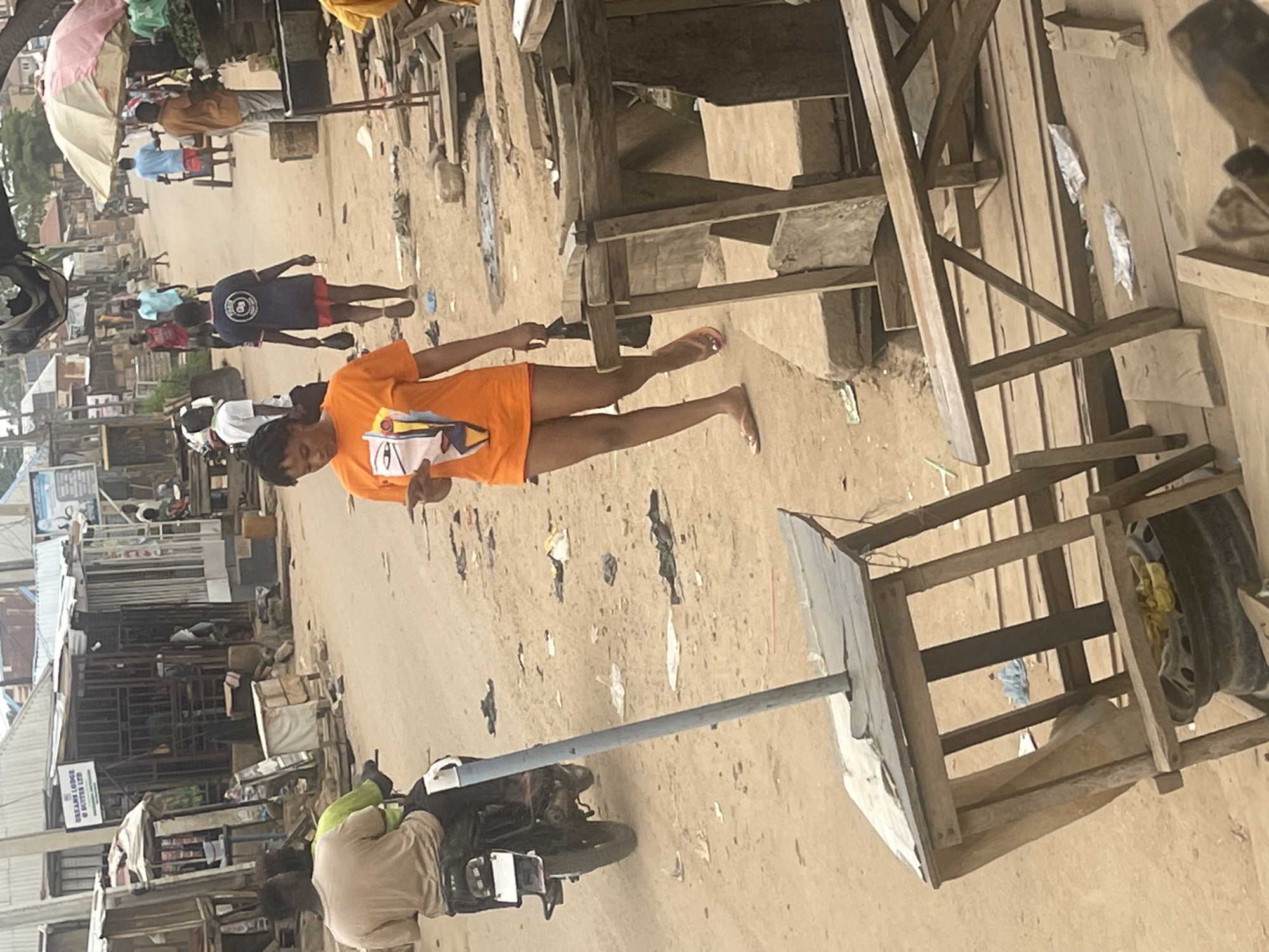 The image depicts a street scene in Abuja, Nigeria, characterized by a bustling marketplace or roadside with various stalls and structures. The ground is sandy and appears dry, suggesting a sunny day. Several individuals are visible, some engaged in activities while others are stationary.
In the foreground, a person wearing an orange t-shirt with a graphic print is standing. Their body is angled towards the right side of the frame, and they appear to be walking or posing. Further in the background, other people are seen, including individuals in blue shirts and red shorts, and someone in a blue t-shirt with a circular emblem. There is a person on a motorcycle, wearing a light-colored garment and a green head covering.
The environment is filled with wooden structures, some resembling market stalls or makeshift shelters, and there are also what appear to be metal frames and scattered debris. A large umbrella, possibly for shade, is visible in the upper left of the image. A sign is partially visible on a building in the background with text that includes "DREAM LODGE & SUCCESS LODGE". The time of day appears to be daytime, given the lighting. The overall atmosphere suggests a lively and somewhat unorganized commercial area.