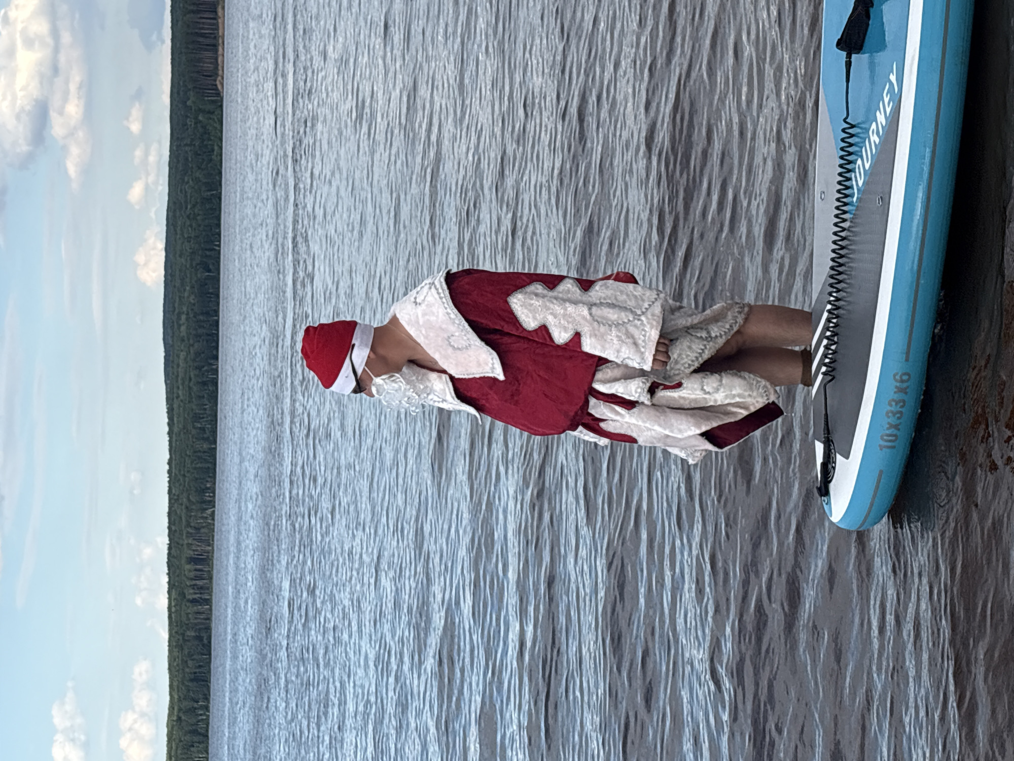 A person dressed as Santa Claus is lying on a paddleboard on a body of water. The paddleboard is blue and white with the word "JOURNEY" printed on the side, along with "10X33X6". The person is wearing a red Santa hat, a red suit with white trim, and a white beard. They are positioned lengthwise on the paddleboard, with their head towards the left side of the frame. The water appears calm with gentle ripples, and the sky is partly cloudy. In the background, a dense green forest line stretches across the horizon. The image seems to be taken during daylight hours, with the lighting suggesting it could be late morning or afternoon. The scene evokes a sense of fun and unexpectedness, as Santa Claus is not typically associated with paddleboarding. The location is identified as Chaikovsky, Russia.