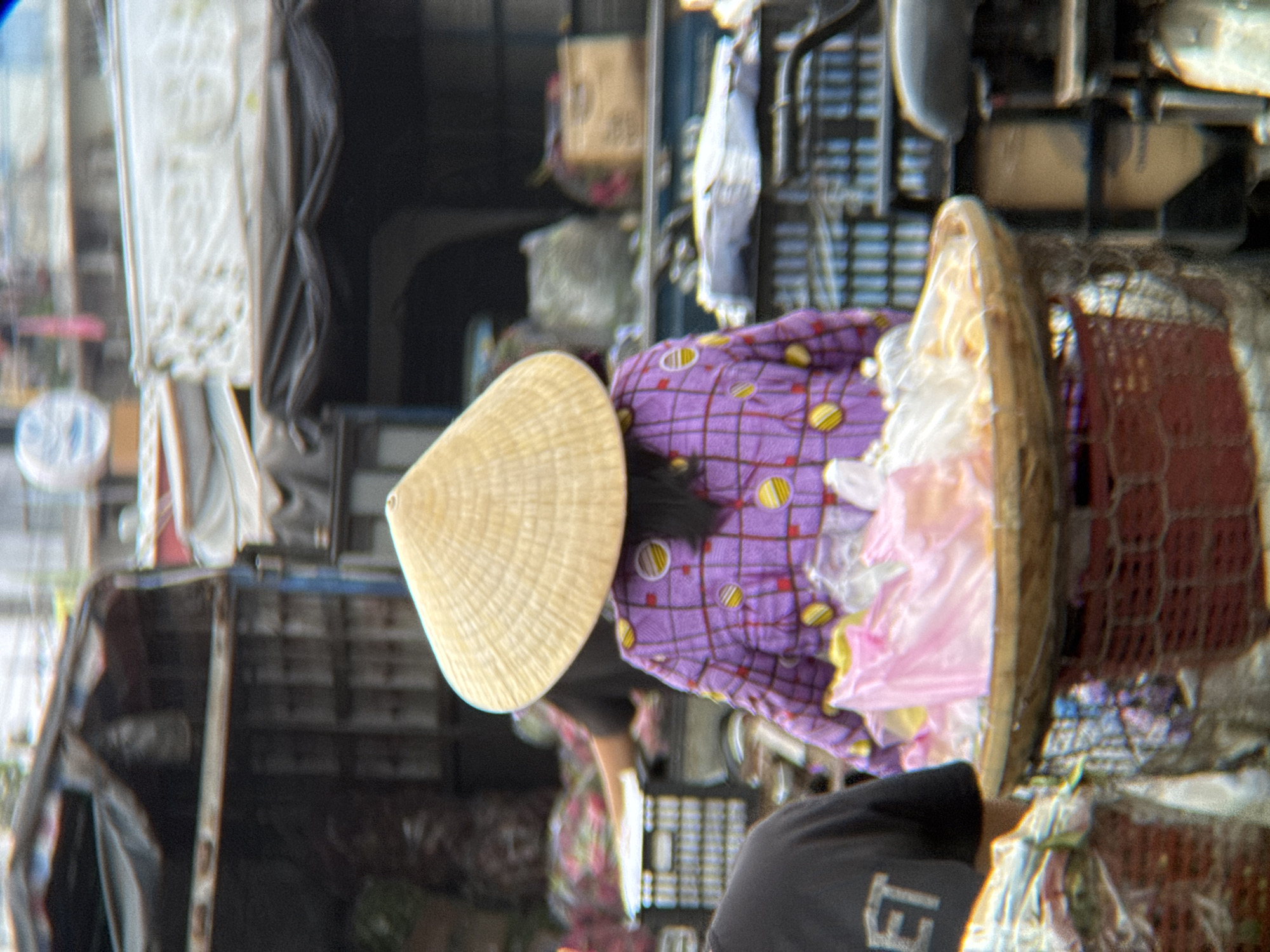 The image depicts a street scene in Nha Trang, Vietnam, likely at a market. A person, viewed from behind, wears a conical straw hat and a purple checkered shirt adorned with yellow circular patterns. They are standing next to a basket filled with pink and white plastic-wrapped items, possibly food or goods for sale, which rests on a red woven basket. To the left, another person, also viewed from behind, wears a black t-shirt with some lettering visible. In the background, there are stalls or vehicles with tarpaulin coverings, crates, and various items that suggest a bustling marketplace. The environment appears to be outdoors, with hints of buildings and sky in the distance, but the focus remains on the foreground figures and their immediate surroundings.