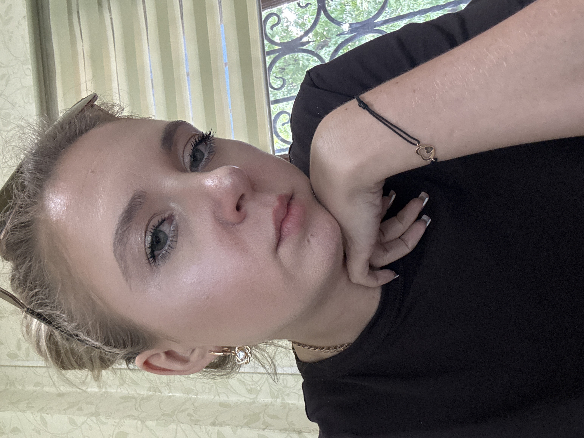 This is a close-up, slightly angled selfie of a young woman. She is wearing a black t-shirt and has her right hand resting on her chin, with her fingers framing her mouth. Her left arm is bent, with her wrist adorned by a thin black bracelet with a small, gold heart charm. She is also wearing a gold necklace and a flower-shaped earring with a clear stone in her visible left ear. Her hair is pulled back and appears to be light blonde. The background features vertical blinds with a subtle floral pattern on the left and a window with decorative black metalwork and foliage visible on the right. The lighting suggests it is daytime. The woman's expression is neutral, and she is looking towards the camera.
