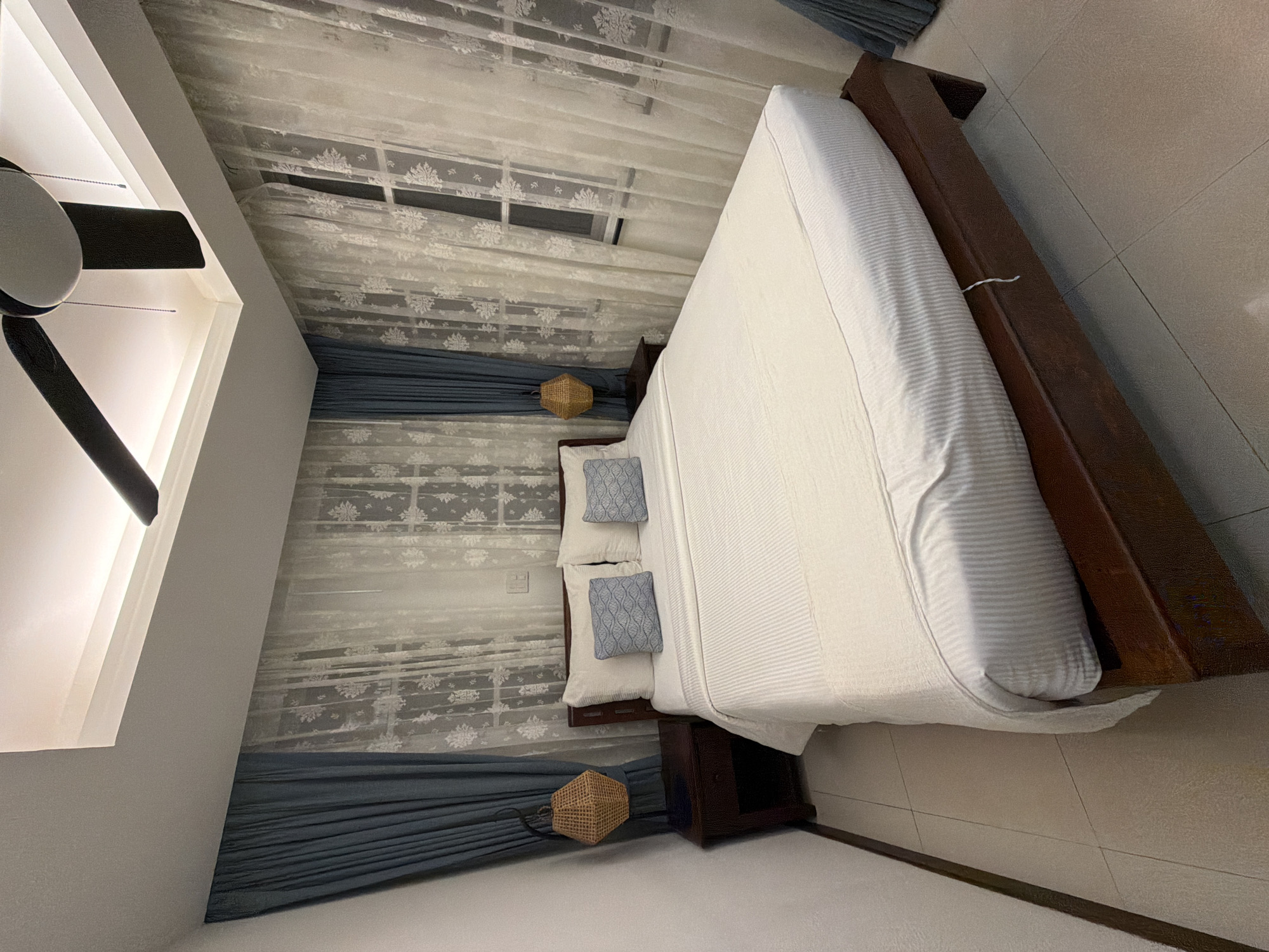 This is a forensic photograph of a well-lit bedroom in Ao Phai, Thailand. The central subject is a queen-sized or king-sized bed with a dark brown wooden frame and a matching headboard. The bed is furnished with white, horizontally striped bedding, including a fitted sheet, a flat sheet, and a bedspread. Two large white pillows are positioned against the headboard, flanked by two smaller decorative pillows featuring a blue and white wavy pattern.
To the left and right of the bed are identical dark brown wooden nightstands, each equipped with a single drawer and an open lower shelf. Suspended above each nightstand is a woven hexagonal-shaped lamp shade emitting a warm light, affixed to the wall with a dark metal fixture.
The wall behind the bed is dominated by windows covered with two layers of curtains. The inner layer consists of sheer white fabric with a floral or snowflake-like pattern. The outer layer comprises solid blue-grey drapes, which are pulled back to frame the sheer curtains. A white electrical outlet or light switch panel is visible on the wall to the right of the bed, between two window sections.
The ceiling is white and features recessed cove lighting that emits a warm, indirect glow around the room's perimeter. A black and white three-bladed ceiling fan is mounted centrally on the ceiling, with pull chains visible. The floor is covered with light-colored square tiles. The walls are painted white. No individuals or animals are present in the scene.