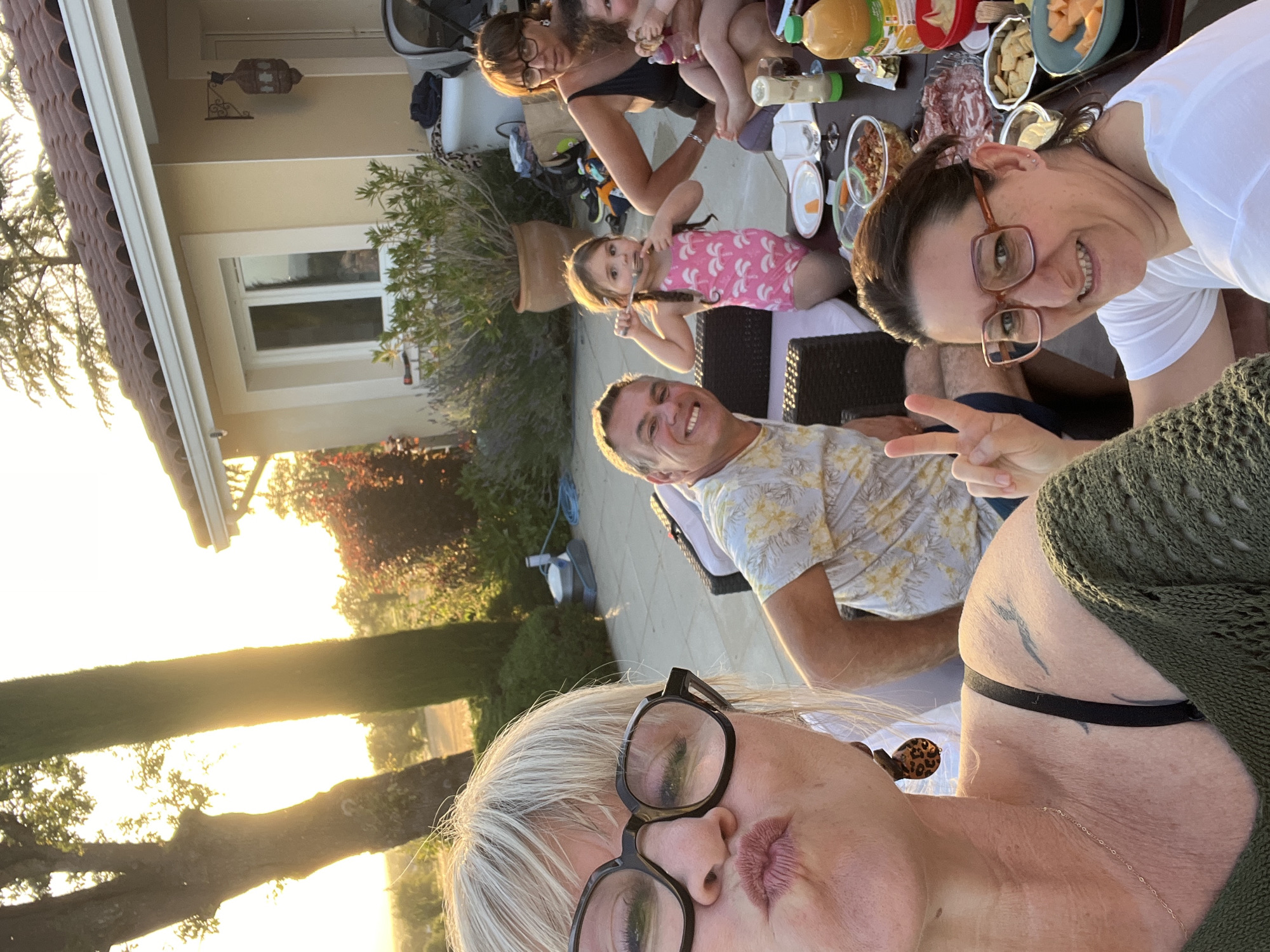 In Saint-Galmier, France, an outdoor gathering takes place in a residential backyard at sunset. Three adults and two young children are present, seated around a table laden with food and drinks. The foreground features a woman with blonde hair and glasses, taking a selfie with a puckered expression, wearing a green knit sweater. To her right, a man with short hair smiles at the camera, wearing a floral shirt. In the background, another woman with dark hair and glasses is seated, holding a baby. A young girl in a pink swimsuit is seen eating with a fork. The environment includes a house with a tiled roof, manicured bushes, trees, and a swimming pool cleaning robot. The warm sunset light illuminates the scene, casting long shadows.