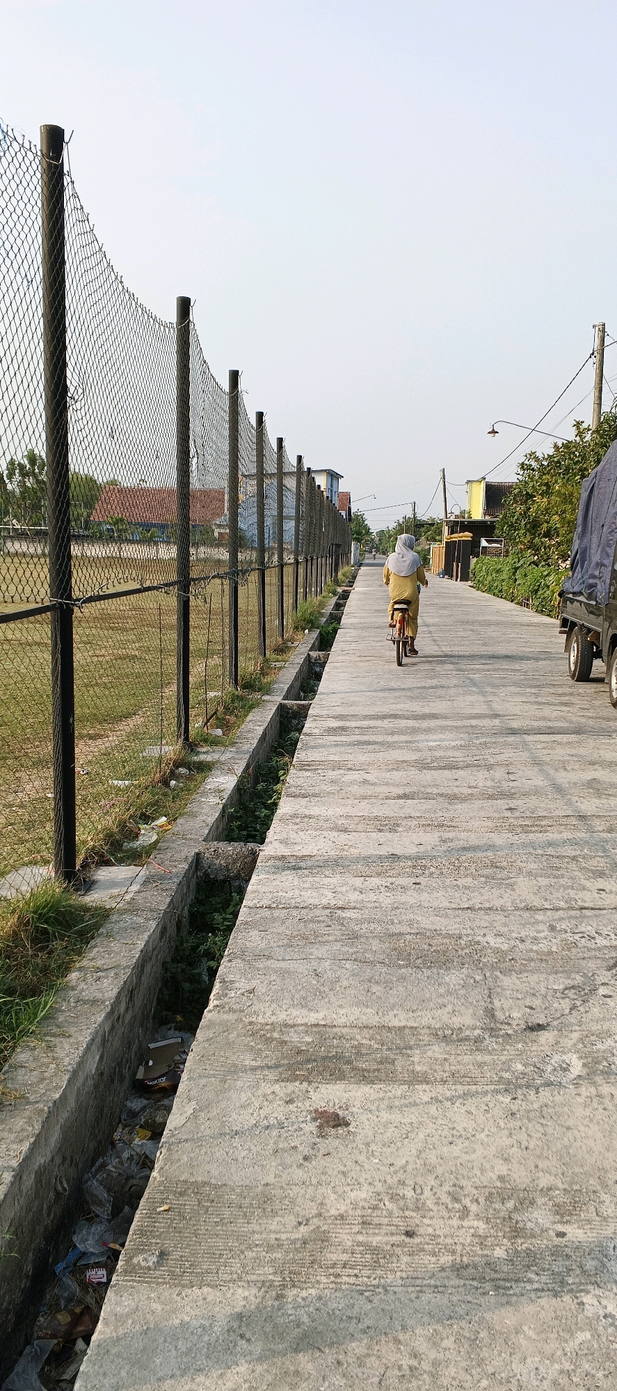 The image shows a person on a bicycle traveling down a paved road. The person is wearing a light yellow garment and a headscarf, obscuring their face. They are cycling away from the camera. To the left of the road, there is a chain-link fence with metal posts, behind which is a grassy field. A concrete drainage ditch runs alongside the road on the left, filled with debris and green vegetation. To the right of the road, there is a partially visible large truck with a covered bed. Further down the road, there are buildings, trees, and utility poles. The sky is clear and pale. The scene appears to be an outdoor area in a town or city, possibly in Latukan, Indonesia, given the context. The time of day appears to be late afternoon or early evening due to the soft lighting.