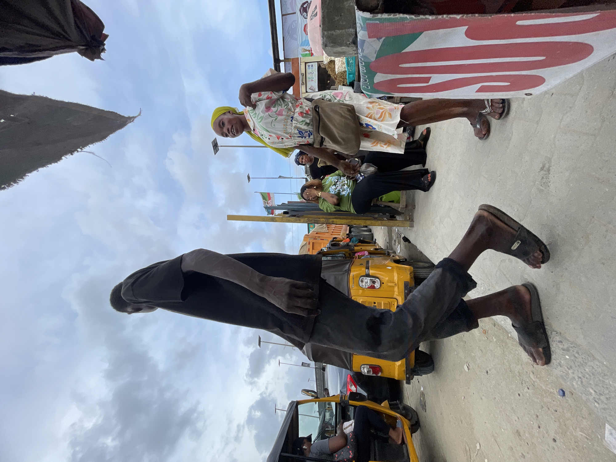 The image captures a vibrant street scene in Lagos, Nigeria, under an overcast sky. In the foreground, a man wearing a dark t-shirt, dark trousers, and black slide sandals is seen from behind, walking towards the right side of the frame, his head lowered. In the mid-ground to the right, a woman stands prominently, smiling towards the camera. She wears a cheerful floral-patterned white dress, a bright yellow headscarf, and flip-flops, with a small brown bag slung across her body. Behind her, another woman is seated, dressed in a green t-shirt and black pants.
The background features several yellow auto-rickshaws, commonly known as Keke Napep, indicating a busy urban environment. Overhead, partial views of market stalls or shaded structures are visible. A large red and white sign with bold lettering, possibly spelling out "PRESS" or similar, is noticeable in the upper right. Further in the distance, a flag with green and white stripes, resembling the Nigerian national flag, is visible. The ground is a light-colored pavement. The scene depicts everyday activity in a bustling city market or street during the daytime.