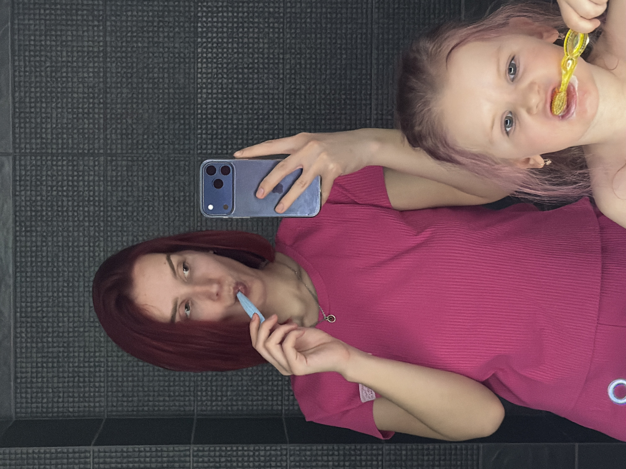 A young woman and a toddler are photographed in a bathroom. The woman is holding a blue smartphone in her left hand while brushing her teeth with a blue toothbrush. She is wearing a pink outfit and a silver necklace. She has auburn hair. The toddler is brushing her teeth with a yellow toothbrush. The toddler has blonde hair with pink ends. The background shows gray tiled wall. The location is Russia, Chaikovsky.