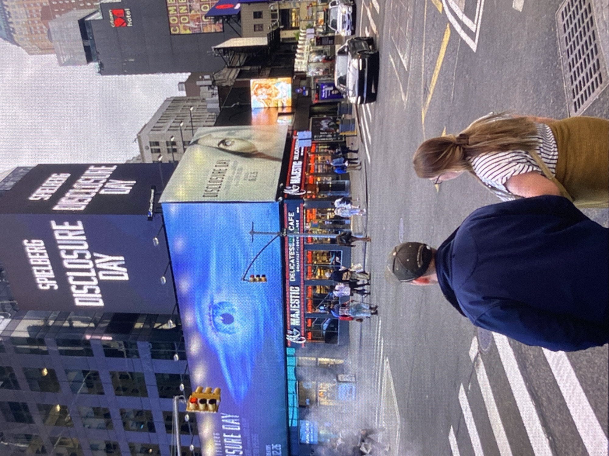 The image shows a street scene in what appears to be Times Square, New York City. The scene is filled with large digital billboards. One prominent billboard displays "Spielberg Disclosure Day" alongside an image resembling an eye in a swirling blue vortex. Other billboards show advertisements and store signs. Pedestrians are walking on the sidewalk. Two individuals are in the foreground, seen from the back, a man in a navy jacket and baseball cap, and a woman with brown hair in a ponytail wearing a striped shirt. Cars can be seen driving on the street. There are traffic lights and crosswalks visible.