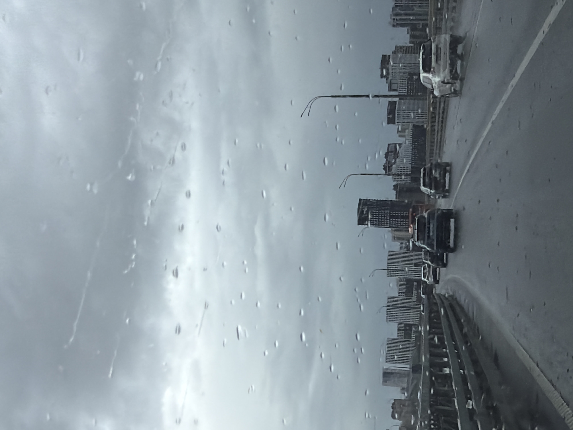 The image depicts a rainy day scene, likely taken from inside a vehicle, looking out at a cityscape. Raindrops are visible on the glass. The buildings on the right appear to be modern, high-rise apartments or offices. Traffic is present on a multi-lane road, with several cars and a larger truck visible. There are lamp posts along the road. The scene could be within the city of Kazan, Russia, given the architectural style of the buildings, but a specific location is not definitively identifiable from this image. The weather is overcast and grey.