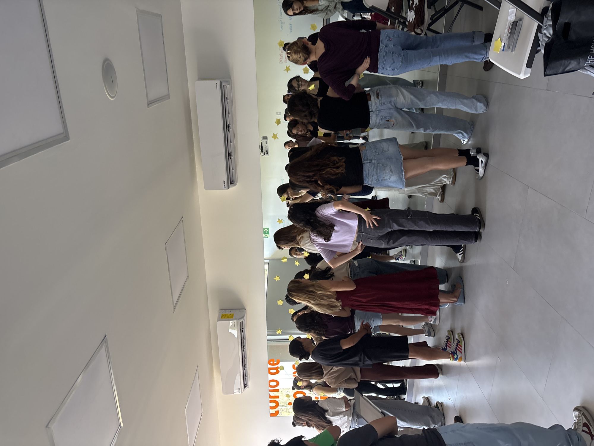 The image captures a group of people, primarily young adults, standing in an indoor setting, likely a classroom, workshop, or training room. The space is modern, featuring white walls, a light-colored tiled floor, and a white ceiling equipped with rectangular recessed light panels and a circular speaker or smoke detector. A white York-brand air conditioning unit is mounted on one wall.
The subjects are arranged in rows, facing a reflective wall that appears to be a large mirror or a glossy whiteboard. Their reflections are visible on this surface. Most individuals are dressed in casual clothing, including jeans, t-shirts, and skirts. Their faces are largely obscured as they are either looking down, facing away, or partially hidden by others. The group's posture suggests they are participating in a structured activity, possibly listening to instructions, observing something, or engaging in a guided exercise.
Notable details include several yellow star-shaped cutouts taped to the reflective wall, and faint, indistinct writings or drawings on its surface, indicating an interactive or creative session. Partially visible text on the wall reads "torio de," which could be part of "Laboratorio de" (Laboratory of) or a similar phrase, hinting at the nature of the facility. An upward-pointing arrow is also visible on the wall. On the right side of the image, a white table with some papers or sticky notes and a dark bag are partially visible. The overall atmosphere appears calm and focused. The location context places this scene in San Andrés Cholula, Mexico.
