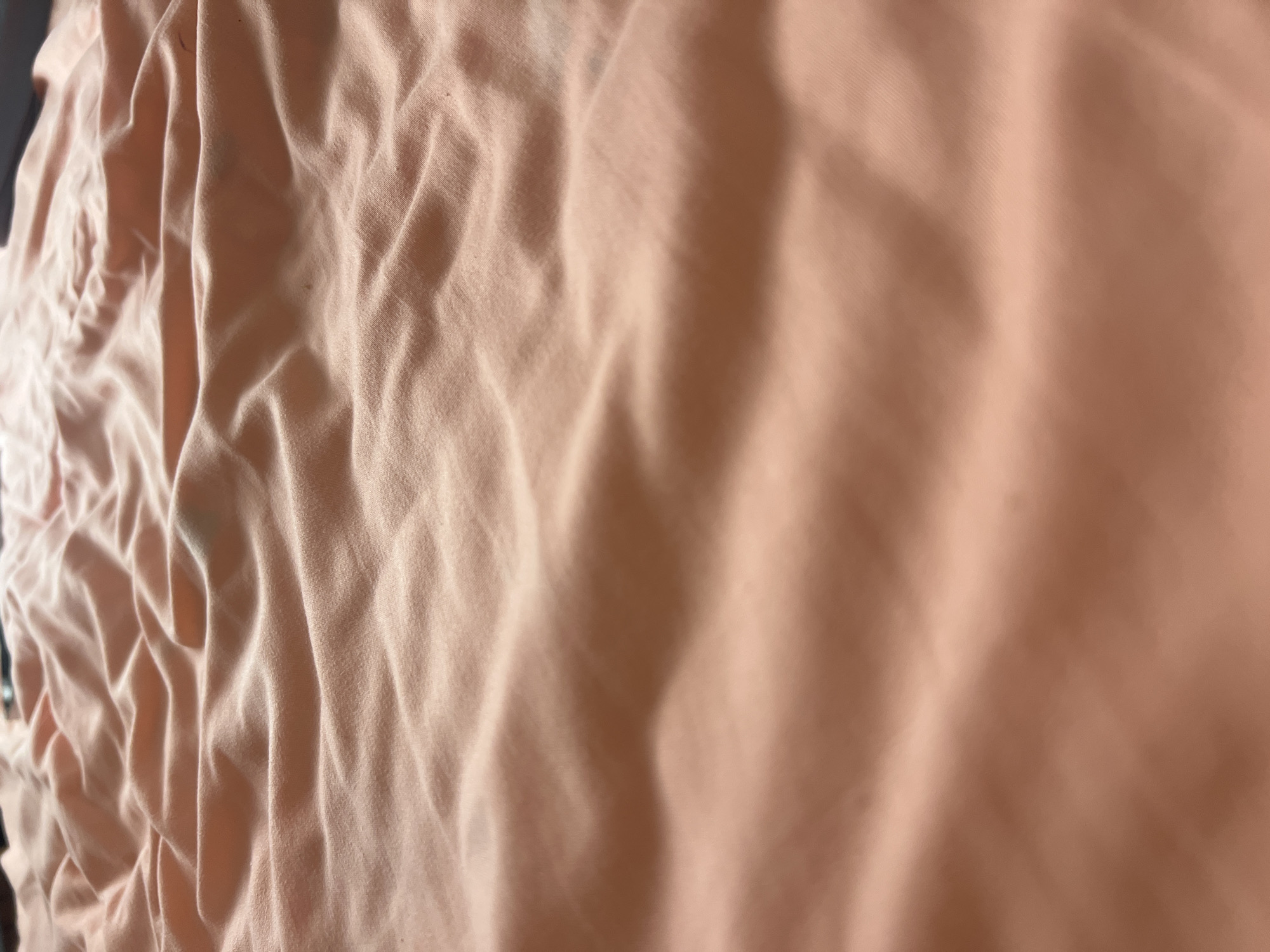The image shows a close-up view of wrinkled peach-colored fabric, likely a blanket or sheet. The fabric is textured with soft folds and shadows that create a sense of depth and movement. The lighting is gentle, highlighting the contours of the material. There are no people or distinct objects visible in the frame, only the fabric itself. The scene is static and offers no information about its location within a city.