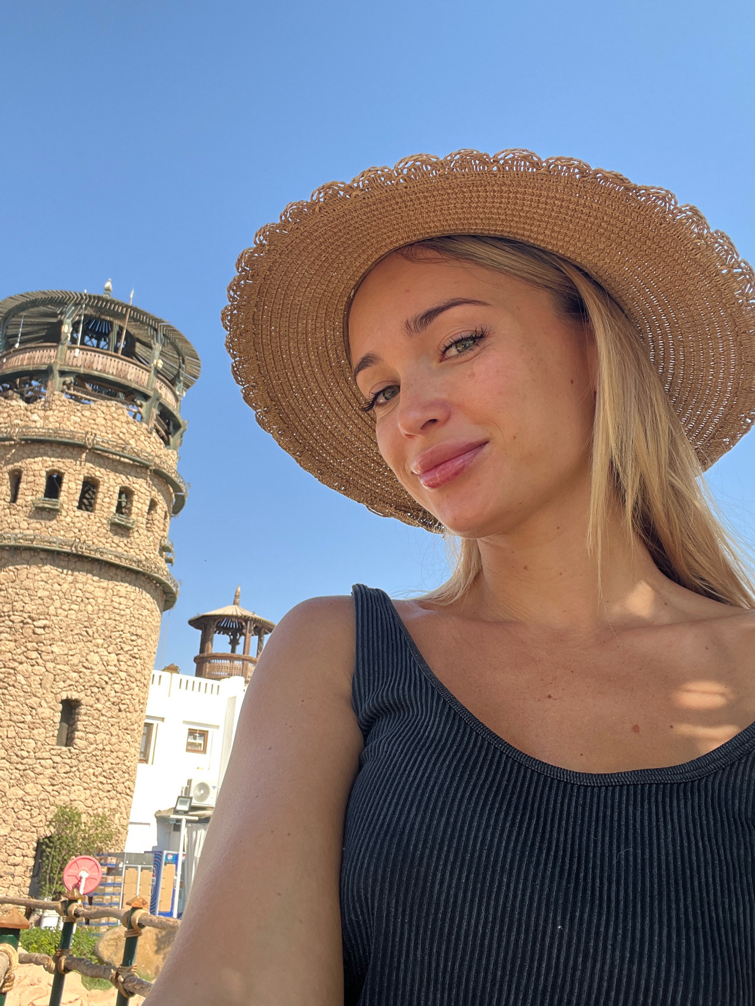 A young Caucasian woman with blonde hair and green eyes is taking a selfie outdoors. She is wearing a wide-brimmed straw hat and a dark ribbed tank top. She is smiling and looking at the camera. In the background, a tall, circular stone tower with multiple levels and balconies is visible. Behind the tower, a smaller, ornate gazebo-like structure can be seen, along with a white building with an air conditioning unit. The sky is clear and blue. The location appears to be in Sharm El Sheikh, Egypt, likely in an area with distinctive architecture.
