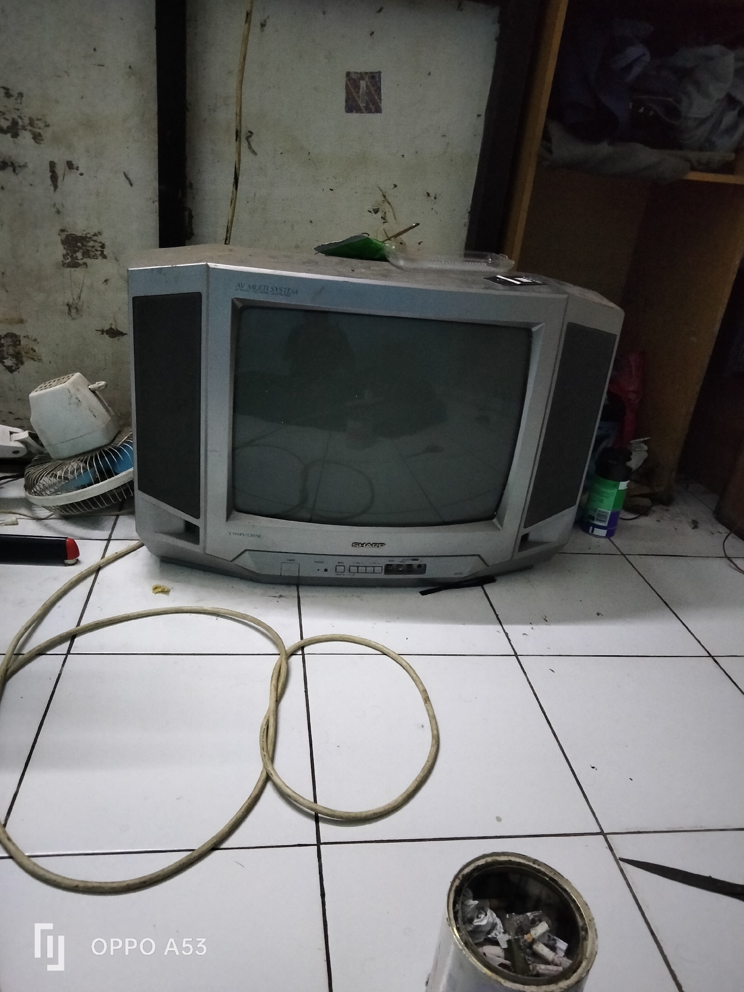 The image depicts an indoor scene, likely a room in Denpasar, Indonesia, with a tiled floor and distressed, grimy walls. Dominating the center is a vintage television set, its silver casing scratched and dusty. The screen is dark. To the left of the television, a small, white electric fan with a blue grill is partially visible, connected to a cord. A thick, coiled power cord lies on the floor in the foreground. To the right of the television, on a shelf, are some clothing items and a can of what appears to be cleaning spray. In the lower right corner, a metal can, possibly an ashtray, is filled with cigarette butts and debris. The overall impression is one of neglect and disarray. There are no people visible in the scene.