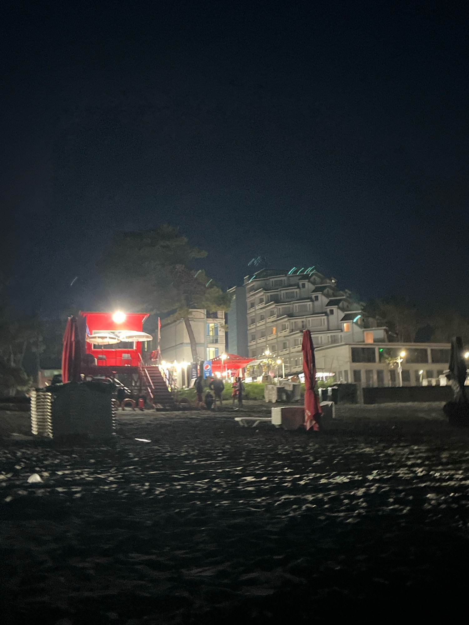 The image shows a beach at night in Ureki, Georgia. The foreground is dark, sandy beach with some scattered debris. In the midground, there is a red structure with stairs leading up to it. This structure appears to be some kind of cafe or bar, with tables and umbrellas visible. Beyond this, there is a large, white, multi-story building with a distinctive terraced design, characteristic of hotels in Ureki. Lights are visible in the windows of the hotel. The sky is dark and clear.