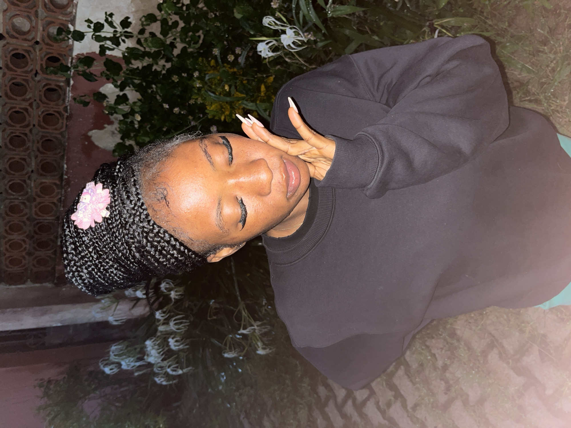 In Jos, Nigeria, a young woman with braided hair adorned with a pink flower is posed outdoors at night. She wears a black sweatshirt and turquoise bottoms. Her eyes are closed, and she is touching her face with her right hand, which has long, pointed nails. The background features plants with white flowers and a textured wall. The ground appears to be paved with bricks and dirt.