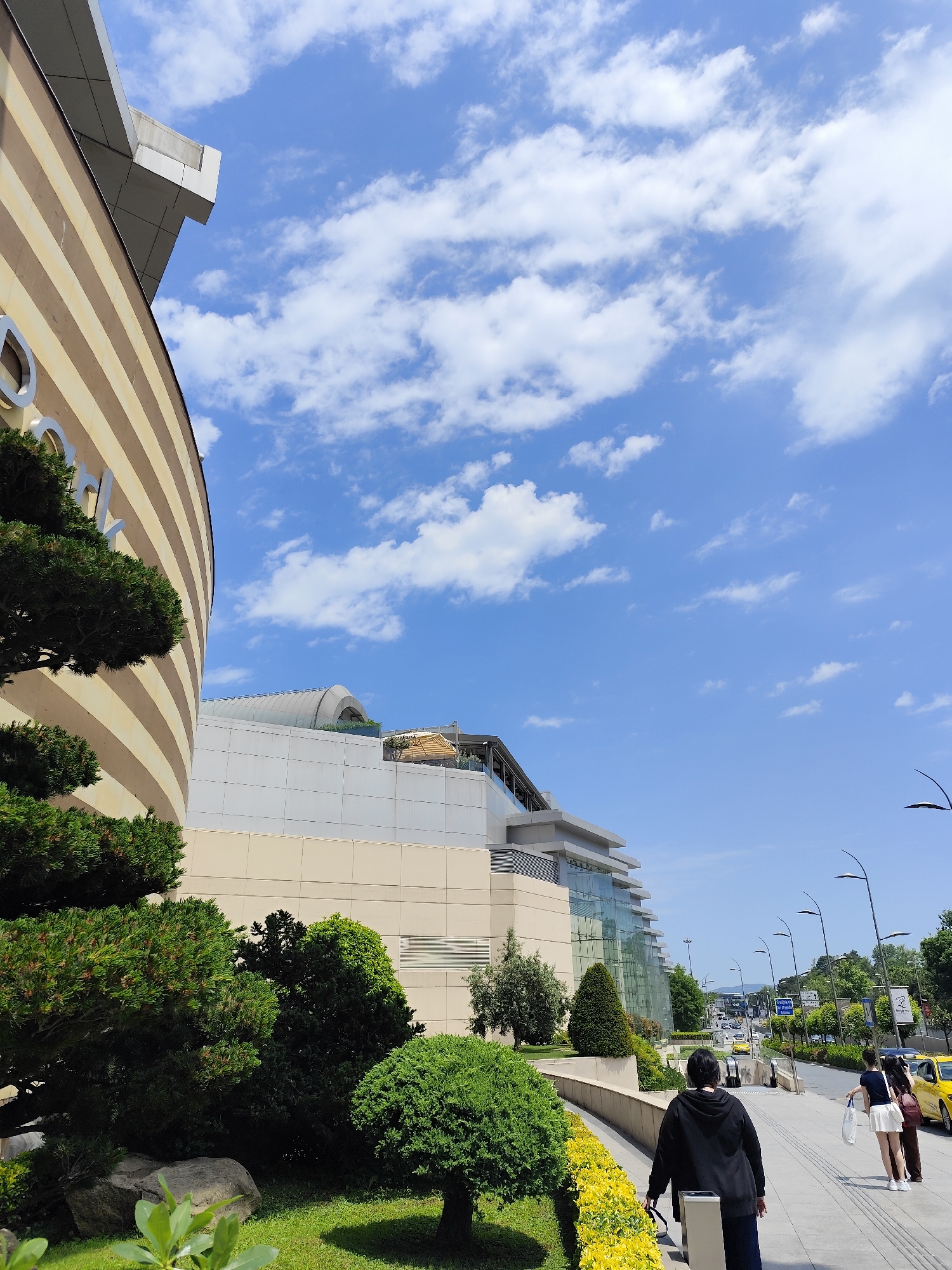 The image captures a sunny day in Sarıyer, Turkey, showcasing a blend of architectural elements and urban life. On the left, a portion of a building with a distinctive horizontally striped pattern is visible, partially obscured by lush, manicured greenery. The building's design hints at a modern or contemporary architectural style.
The foreground features a pathway lined with yellow flowers, where several people are walking. One individual, facing away from the camera, is wearing a black hoodie and dark pants. Further ahead, two women are walking side-by-side, one dressed in a white skirt and the other in dark attire.
Beyond the pathway, a road with light traffic is noticeable. Yellow taxis and other vehicles can be seen, suggesting an active urban environment. Streetlights line the road, and trees add a touch of greenery to the roadside.
The sky is bright blue with scattered white clouds, contributing to the sunny and clear atmosphere of the scene.