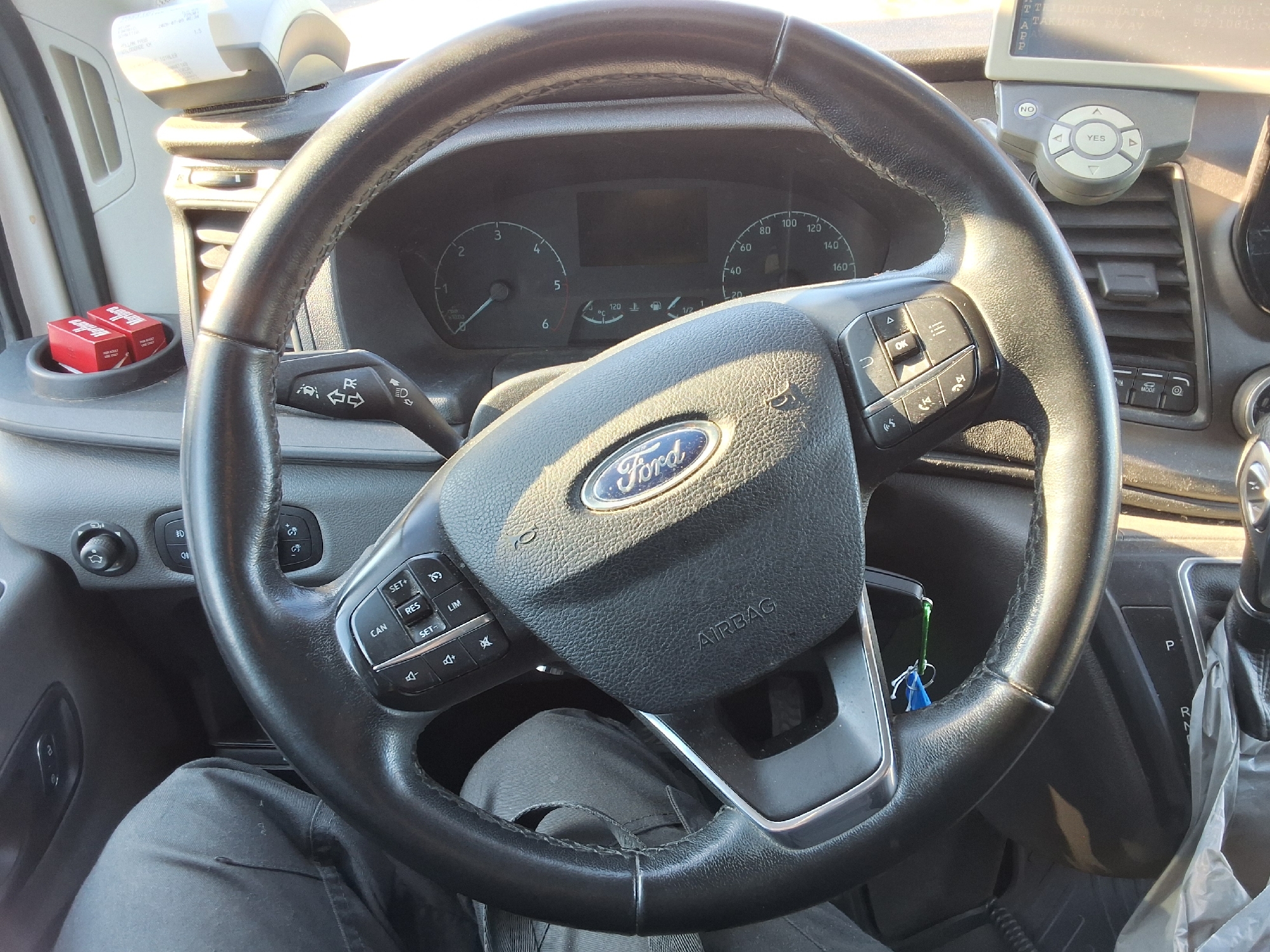 The image shows the interior of a vehicle, likely a van, with a focus on the steering wheel and dashboard. The steering wheel is black leather with a Ford logo in the center. Various buttons and controls are visible on the steering wheel, including cruise control buttons and media controls. The dashboard includes a speedometer, tachometer, and a digital display. To the left of the steering wheel, there is a cup holder containing two red cigarette packs. Above the cup holder, a small device with a receipt is attached to the dashboard. A portion of a person's leg, clad in dark pants, is visible in the lower part of the image, suggesting they are in the driver's seat. The overall environment appears to be indoors, possibly in a parking lot, given the ambient lighting and the presence of a vehicle interior. The location in Arboga, Sweden is not discernible from the visual information provided in the image.