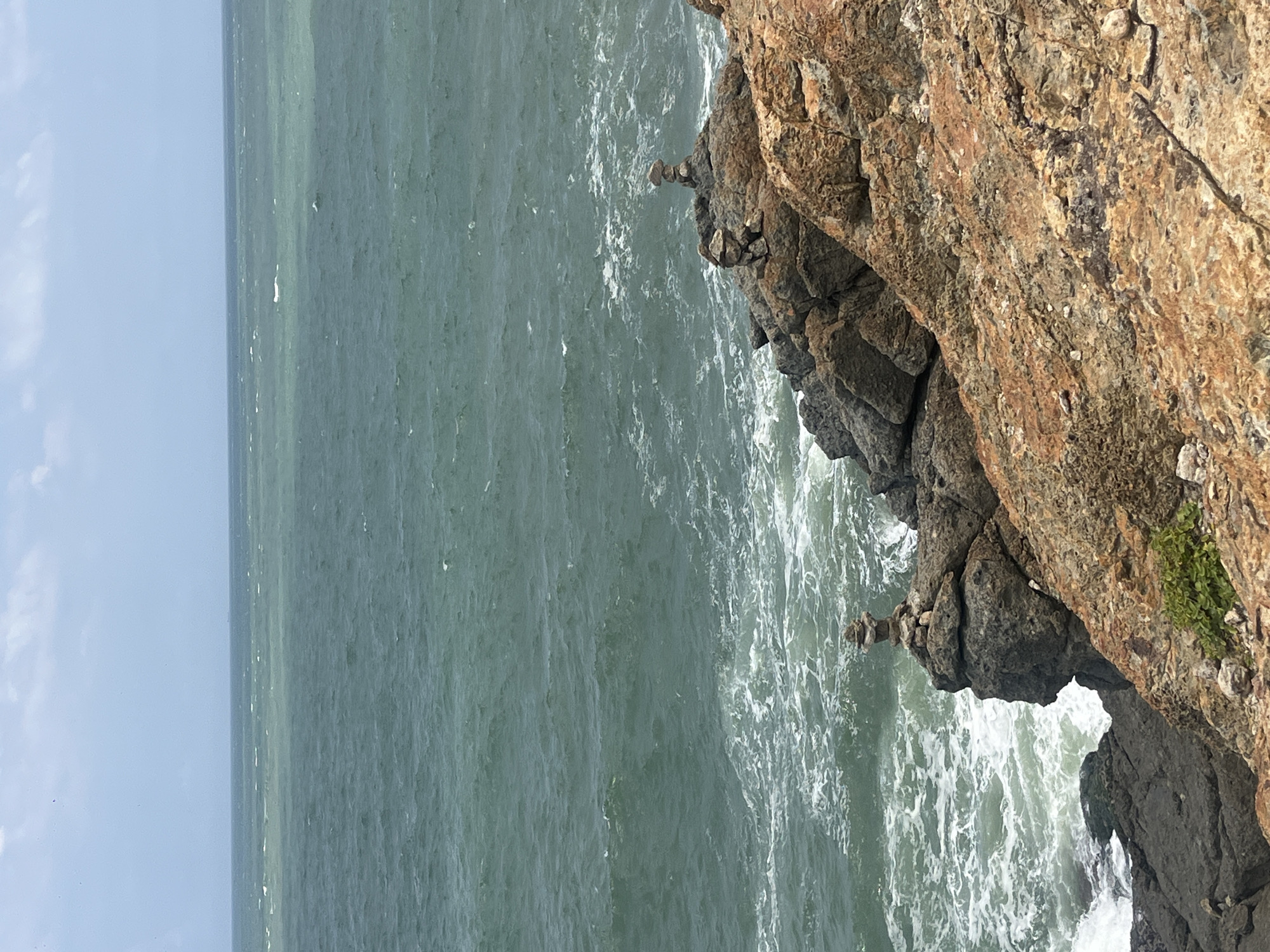 The image shows a rocky coastline with the sea in the background. In the foreground, there are two rock formations, each with a stack of smaller stones balanced on top. The sea is a deep green color with whitecaps, and the sky is a pale blue with a few white clouds. The rocks are varied in color, with shades of grey, brown, and orange. The location is identified as Mui Ne, Vietnam. The scene is still and peaceful, with no people or animals visible.