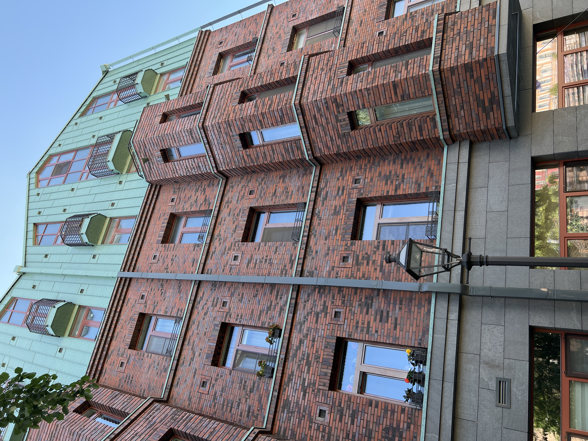 The image captures a close-up, worm's-eye view of the facade of a red-brick building with a distinctive mint-green, multi-pitched roof. The building appears to be located in Saint Petersburg, Russia, given the architectural style and the Cyrillic text visible in the reflection of a lower window.
The facade is constructed with varying shades of red and brown brick, arranged in horizontal courses. Numerous windows with reddish-brown frames punctuate the brickwork. Some of these windows are adorned with small planters containing flowers. The building features protruding sections, including balconies with dark metal railings. A prominent dark grey downspout runs vertically along the brick facade.
Below the brickwork, a section of the building is clad in large, grey stone tiles. In this lower section, a dark, ornate street lamp stands in the foreground, positioned to the right of the downspout. Reflections of buildings and trees are visible in the windows of the lower floors. The sky above is clear and blue. The overall impression is one of detailed, classic architecture with modern elements.