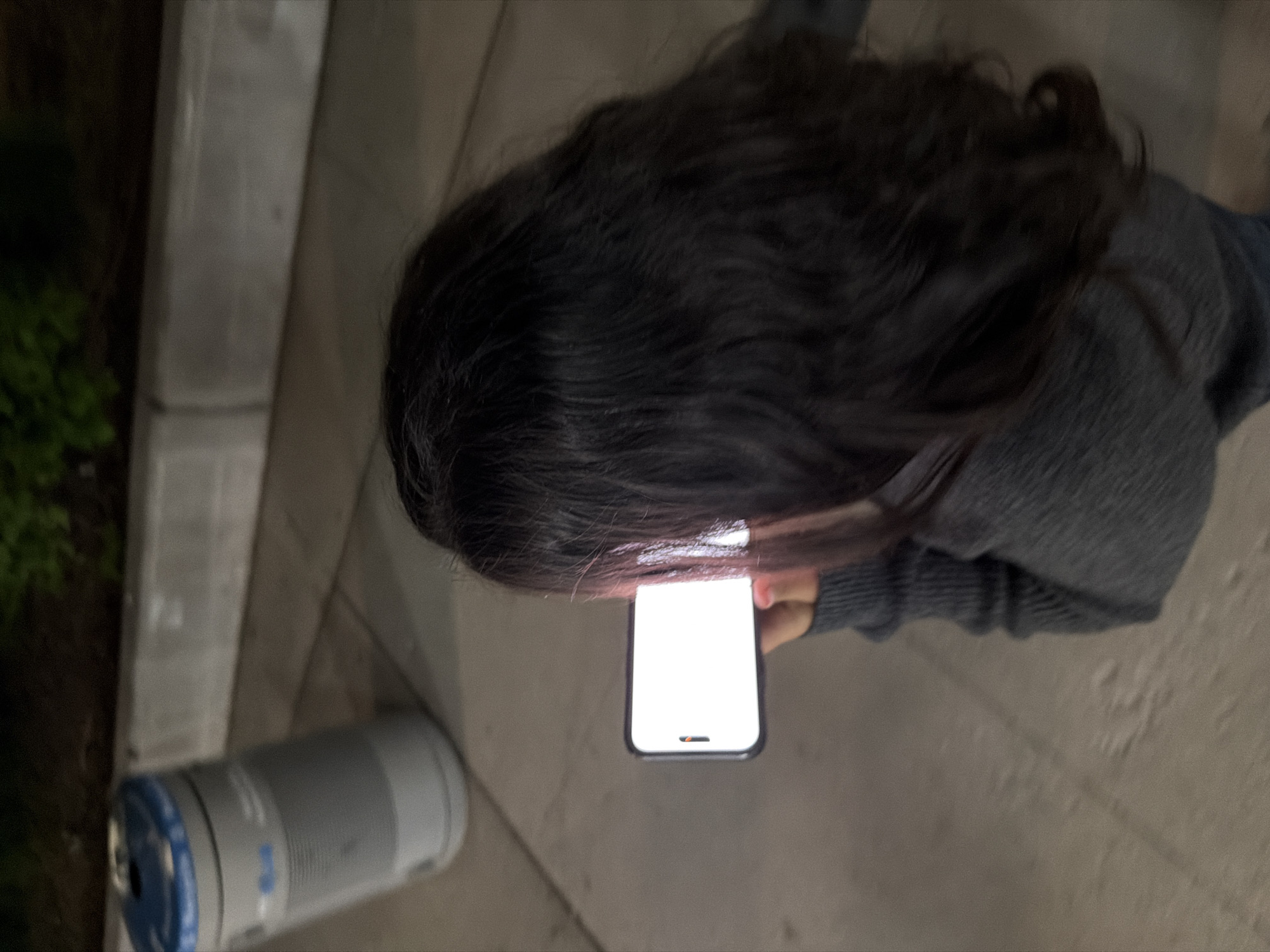 An individual with long, dark hair holds a brightly illuminated mobile phone, likely a smartphone, in an outdoor setting. The person is wearing a gray sweater. The surrounding environment includes what appears to be a concrete sidewalk or pavement and nearby steps. To the left is a cylindrical object, possibly a container or bottle. The location cannot be determined solely from the image. The image does not allow to determine the specific location in the city of New York, United States of America.
