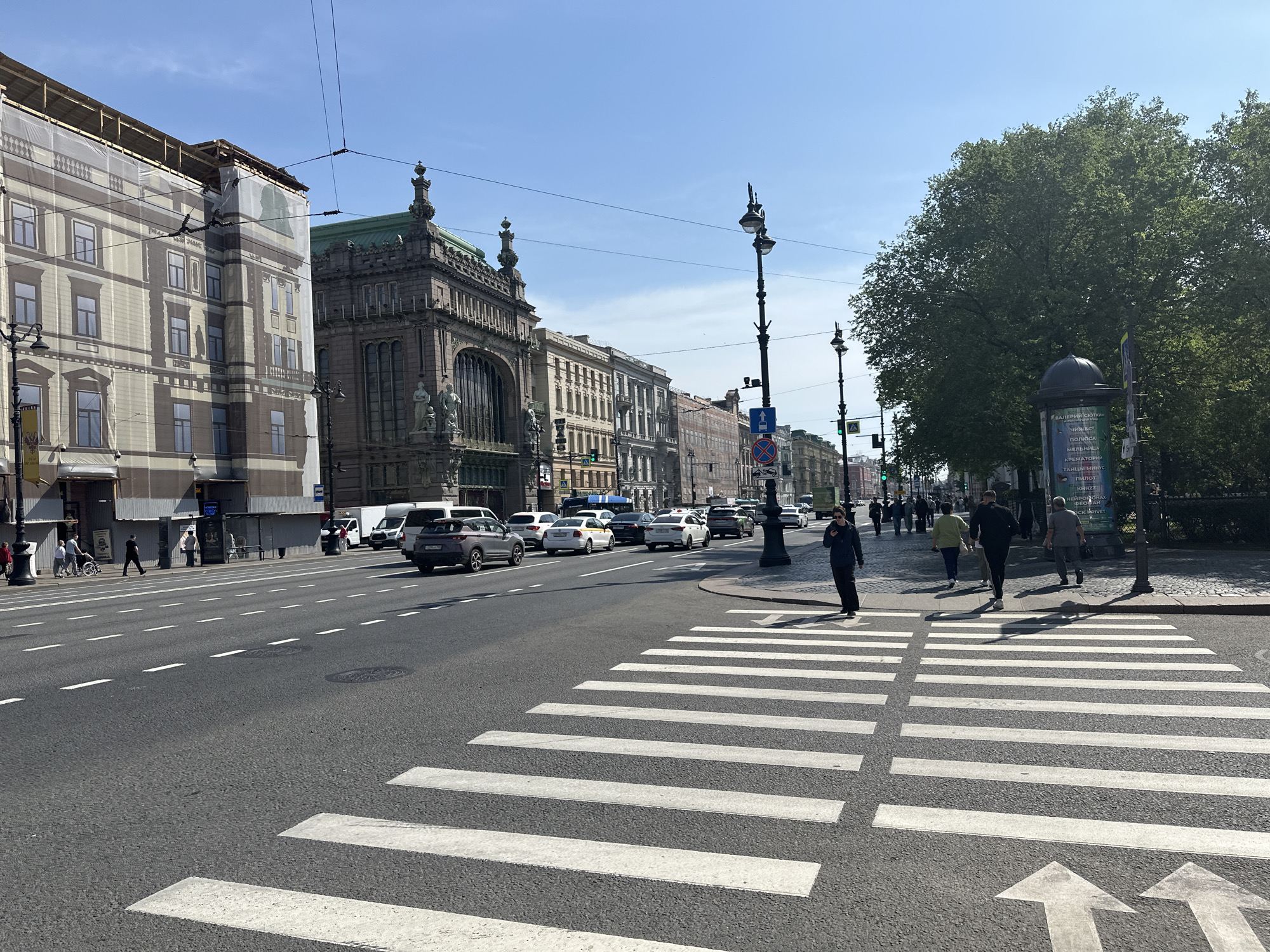 The image depicts a street scene in Saint Petersburg, Russia on a sunny day. A wide, multi-lane road with a crosswalk dominates the foreground. Arrows are painted on the asphalt indicating lane direction. A group of pedestrians are walking across the crosswalk, while others stroll on the sidewalk to the right. Cars, vans, and taxis occupy the road lanes, moving in both directions. The architecture on either side of the street is characterized by ornate, classical buildings. On the left side, a building is partially covered with a large screen that mimics the facade. In the background, a distinct building with green roofing and sculptures stands out. Streetlights and traffic signs are positioned along the sidewalks. The sky is clear blue. Trees with dense green foliage border the right side of the street. A column advertising various performances stands next to the trees. The overall scene is a bustling urban environment.