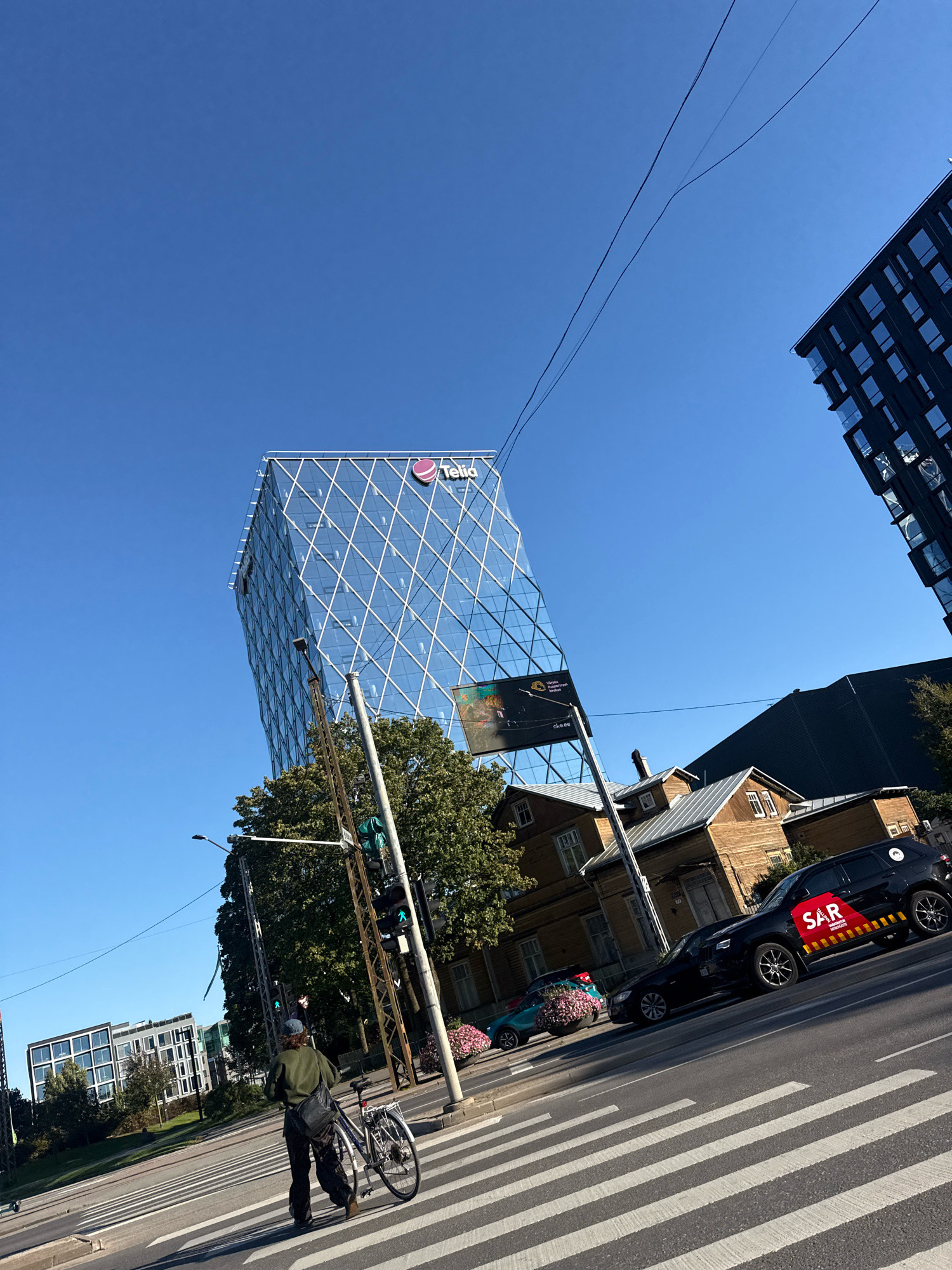 A person pushes a bicycle across a crosswalk in front of a modern glass building with the "Telia" logo. To the right of the glass building are older wooden houses and a black car with a red "SAR" logo. The sky is clear blue, and the sun is shining, casting shadows on the street. In the background, there are more buildings and trees. The image appears to be taken during daylight hours. The location is likely Tallinn, Estonia, as indicated by the "Telia" building, which is a prominent landmark in the city.