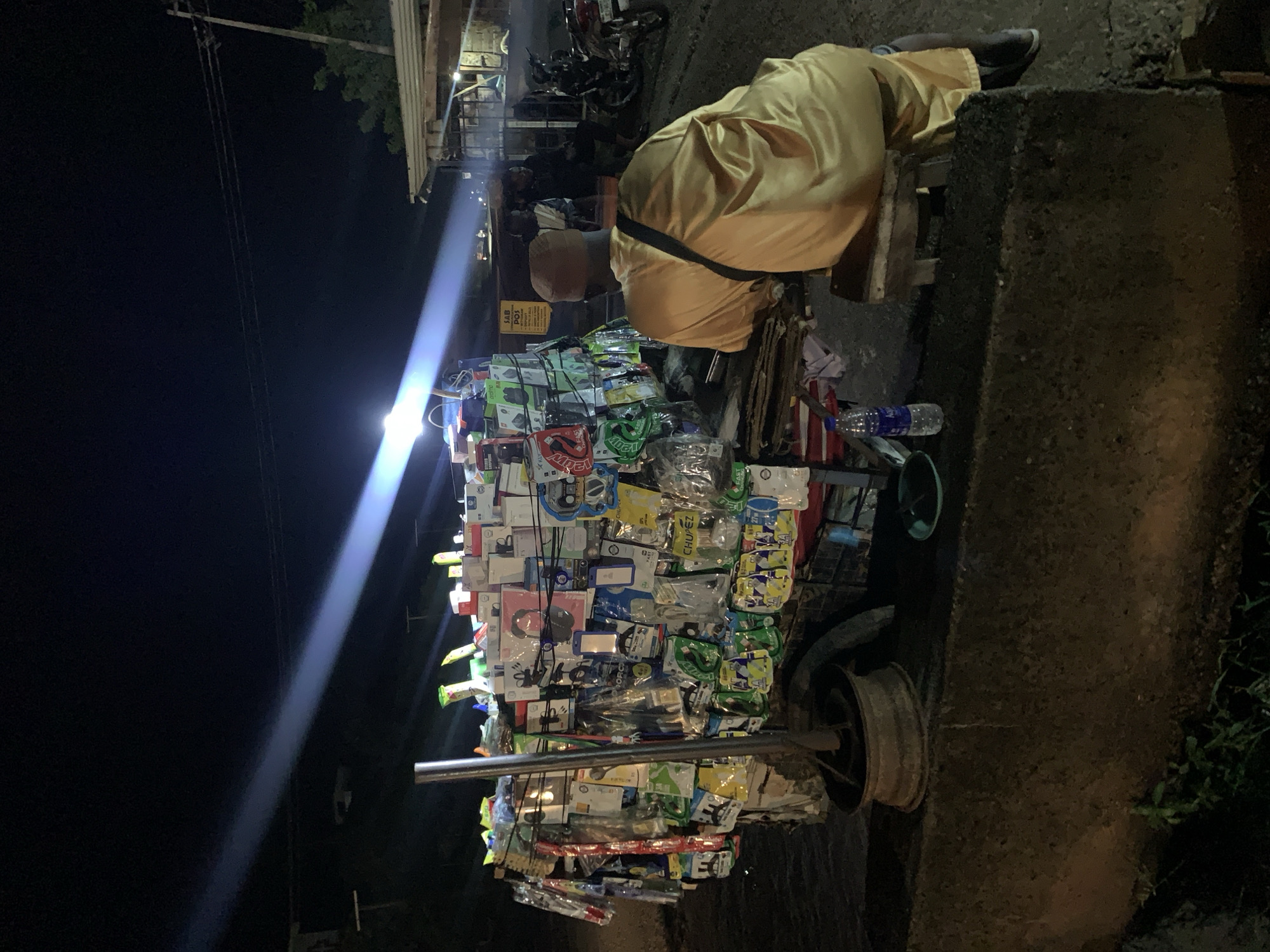 This is a night-time photograph taken on a street in Lokoja, Nigeria. A vendor's cart, laden with an assortment of electronic accessories such as phone cases, chargers, and cables, is illuminated by a bright overhead light, casting a long beam across the dark sky. The cart is positioned on a concrete embankment. In the foreground, a person wearing a mustard-yellow garment and a cap sits on a wooden stool, facing away from the camera, looking towards the cart. In the background, beyond the cart, are structures that appear to be market stalls or shops, and a motorcycle is visible. The scene suggests a commercial street operating at night, likely a roadside market.
