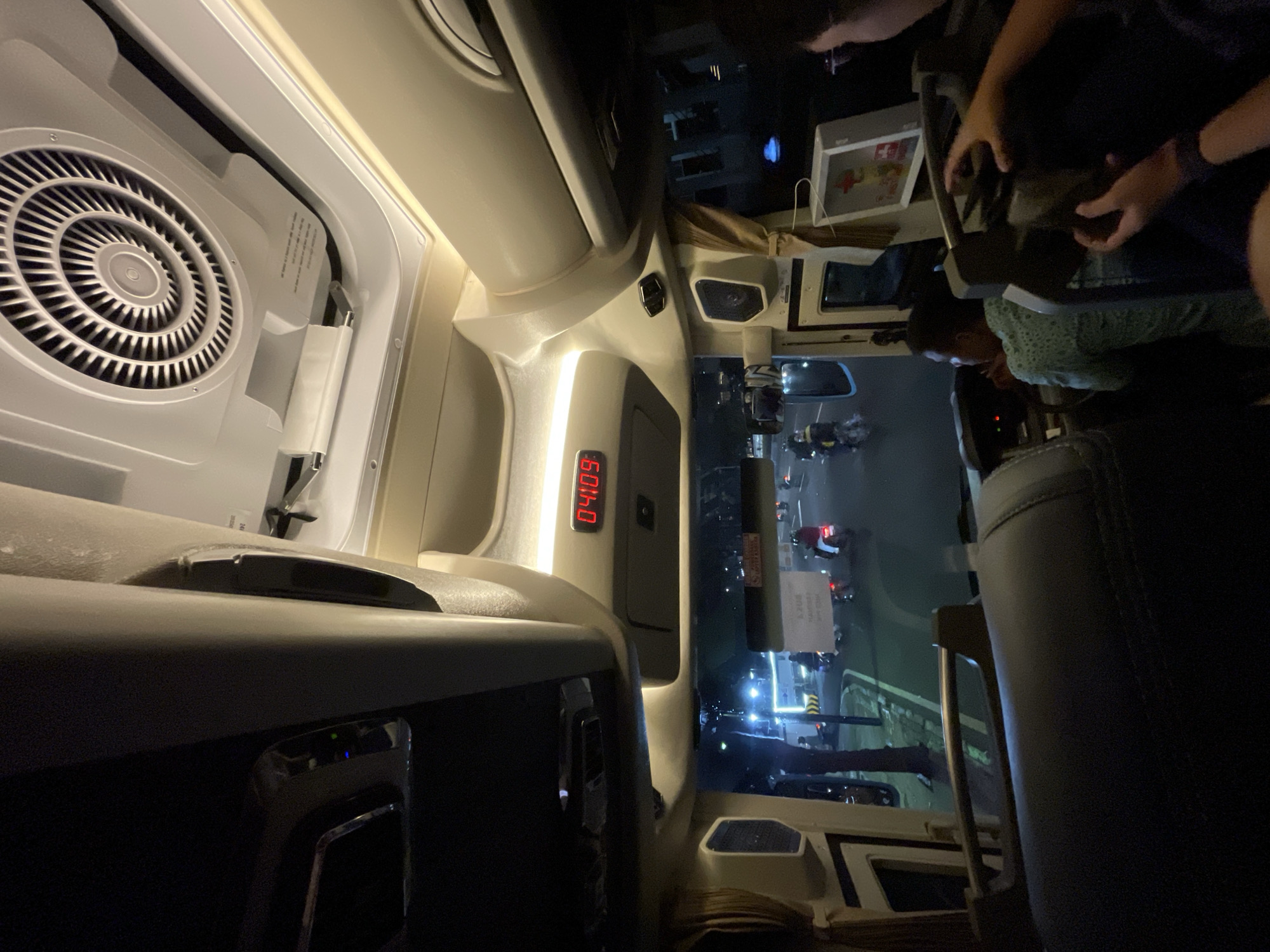 The image shows the interior of a vehicle, likely a van or bus, at night, oriented vertically. The view is forward, through the windshield, revealing a street scene.
**Setting:** The street scene appears to be in Central Jakarta, Indonesia, based on traffic patterns, signage, and overall urban environment. It is nighttime with artificial lighting illuminating the road and surrounding buildings.
**Subjects/Objects:**
* **Vehicle Interior:** The image focuses on the vehicle's interior, showing elements such as the dashboard, air conditioning vent, a digital display showing the time "04:09", and passenger seating.
* **People:** Several people are partially visible. A driver is seen partially in view looking forward. There are passengers behind the driver handling something, possibly food or documents.
* **Street Scene:** Traffic is visible outside, including cars, motorcycles, and streetlights.
* **Objects outside:** An ambulance, a street light, buildings, and vehicles.
**Actions/Interactions:**
* The driver is focusing on driving.
* Passengers are interacting with objects in their laps.
**Environment:**
* The environment is an urban setting at night.
* Lighting conditions inside the vehicle are subdued, with light coming from the dashboard and instrument panels, as well as from outside.