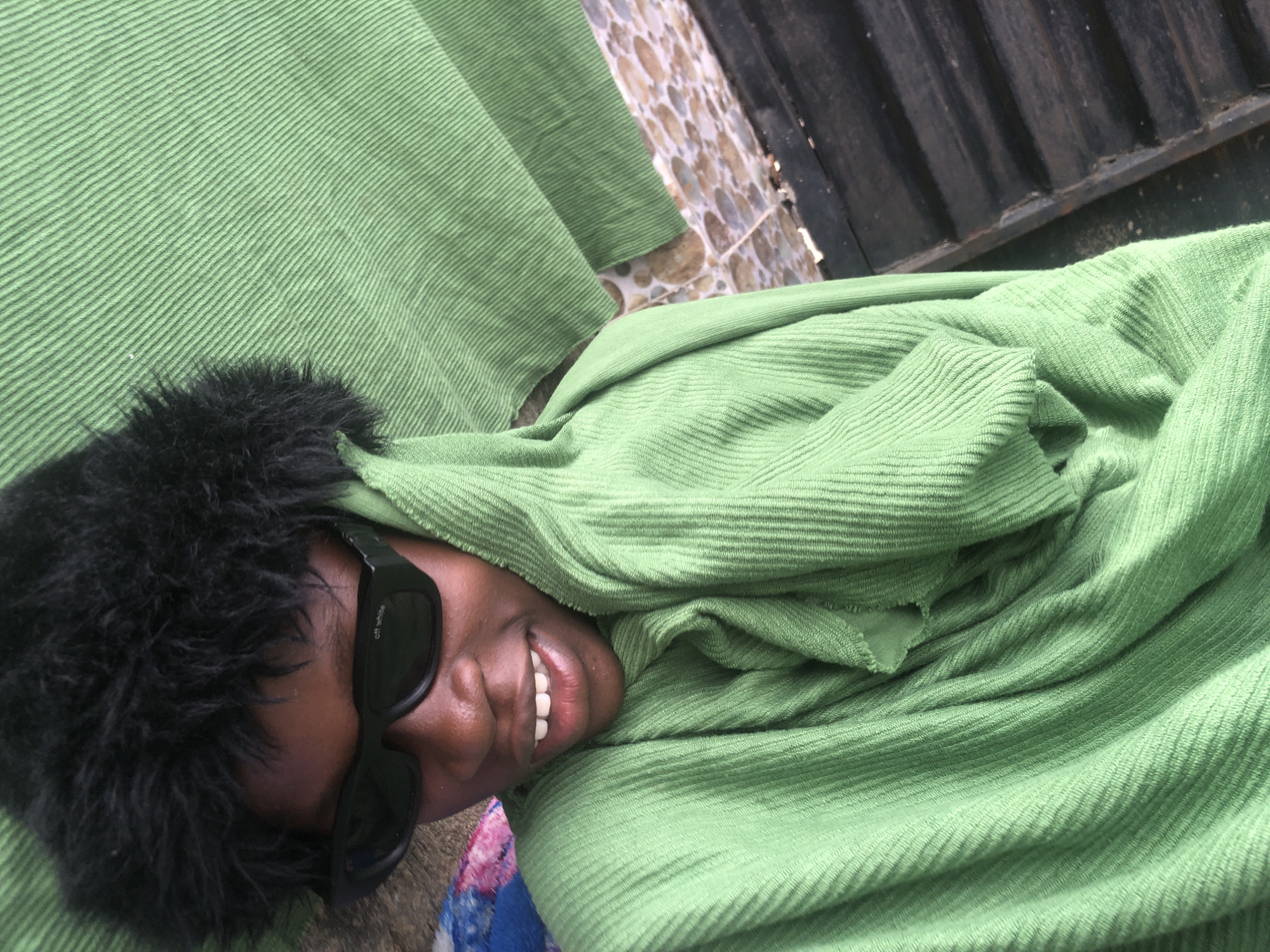 A close-up, high-angle shot shows a young Black person wearing a black fuzzy hat and black sunglasses, draped in a light green towel. The person is smiling broadly, with their head tilted to the side. The towel is wrapped around them, obscuring most of their body. The background is blurred, but appears to be an outdoor setting with a patterned tiled surface and dark wooden planks. The image is likely taken in Ado, Nigeria, given the context provided.