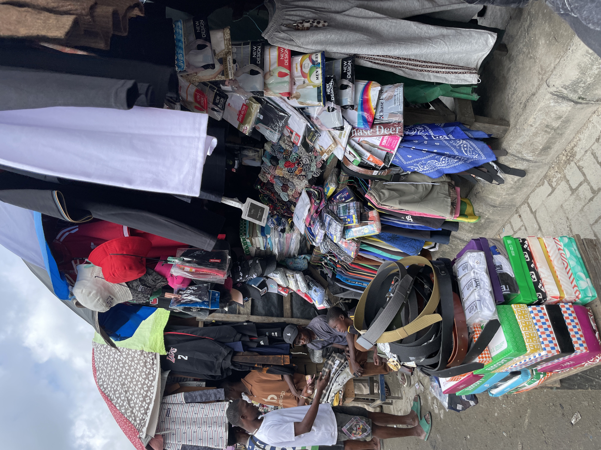 In Lekki, Nigeria, a street vendor displays an array of clothing and accessories under a makeshift canopy. The scene is crowded with various items of apparel, including t-shirts, shirts, pants, belts, and hats. Several men are present, appearing to be either vendors or customers. One man in a white t-shirt and shorts is interacting with another man seated on a stool, possibly making a purchase or discussing goods. Another man in a brown shirt is also seated. The ground is paved with concrete and cobblestones. The overall impression is of a bustling market environment.
