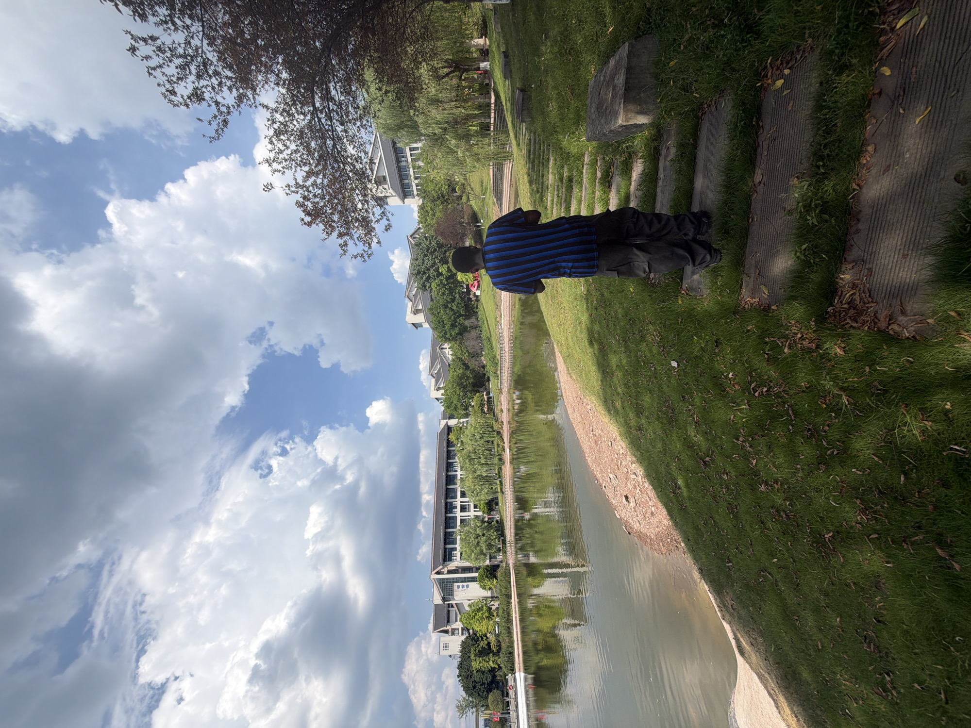 This image depicts a scenic view of a park-like area with a lake in the foreground and buildings in the background, likely located in the city of Jiangjin, China. The sky is partly cloudy with a mix of blue sky and white, fluffy clouds. A person, seen from behind, is standing on a stone pathway covered with grass. The person is wearing a blue and black striped t-shirt and dark, baggy pants. The pathway leads upwards, away from the viewer, towards more greenery and trees. The lake is calm and reflects the surrounding trees and buildings. Along the edge of the lake, there is a narrow strip of sandy or gravelly shore. In the background, several modern buildings with dark roofs are visible, nestled amongst lush green trees and landscaping. The overall atmosphere suggests a pleasant day in a well-maintained outdoor recreational space.