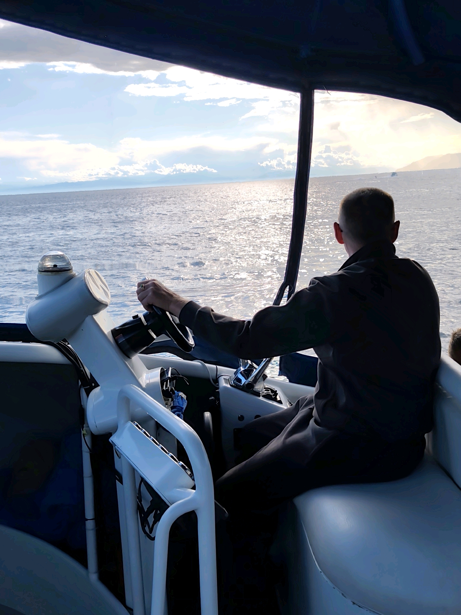 The image shows a person, viewed from behind, steering a boat on a body of water. The person is wearing dark clothing and has short hair. They are seated at the helm, with their hands on the steering wheel. The boat appears to be moving, as indicated by the rippling water. The environment is a large body of water, possibly Lake Baikal, with a distant shoreline visible under a cloudy sky. The sun is shining, creating a shimmering effect on the water. The boat has a white console with various controls and a clear light fixture on top. The image is taken from within the boat, looking out towards the water and the horizon. The location is not specified in the image, but given the context of Listvyanka, Russia, it's likely a scene from this settlement on the shore of Lake Baikal.
