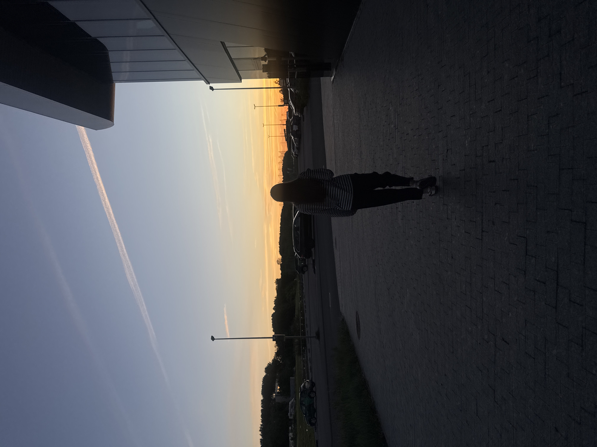 A person with long dark hair walks away from the camera on a paved walkway towards a modern building with dark cladding and glass windows. The person is silhouetted against a colorful sunset sky with a contrail. In the background, there is a parking lot with several cars and a line of trees. The scene appears to be located in a suburban or commercial area, possibly on the outskirts of Tallinn, Estonia.
