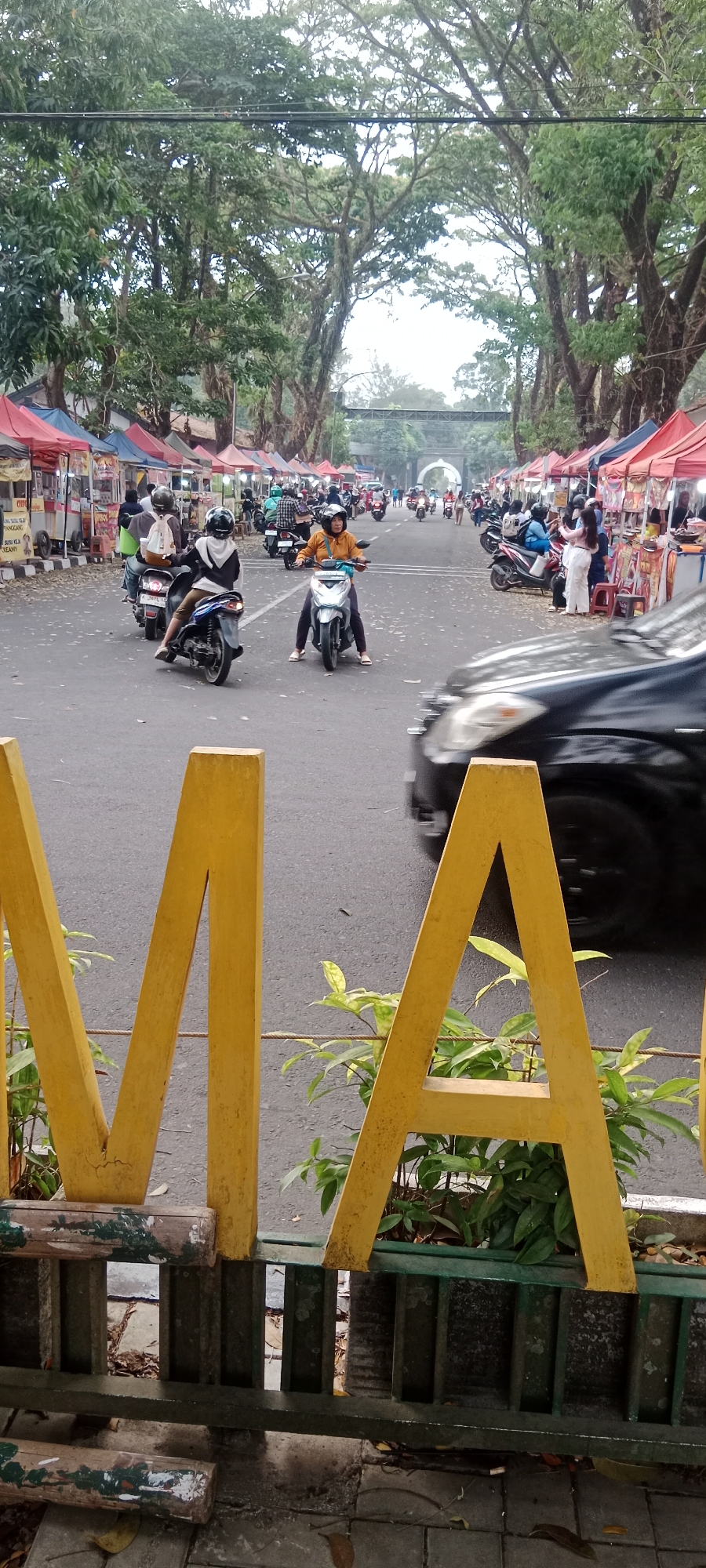 The image shows a street scene in Magelang, Indonesia, likely during the day. The foreground features large, yellow, wooden letters spelling "MA," which are part of a decorative sign. Behind the letters, a street runs through the scene, lined on both sides by rows of market stalls or temporary shops, identifiable by their red and blue canopies. A black car is blurred in motion in the mid-ground. Several people are present, most of them riding motorcycles. One woman in an orange jacket is riding a white scooter, facing forward. Another person wearing a black and white hoodie is riding a blue scooter, facing away from the camera. Other motorcycles and a few pedestrians can be seen further down the street. The street is bordered by large, mature trees with dense foliage, creating a canopy effect over the road. In the distance, an arched gateway is visible. The ground is paved with asphalt and some scattered dry leaves. The overall impression is that of a busy market area along a tree-lined street.
