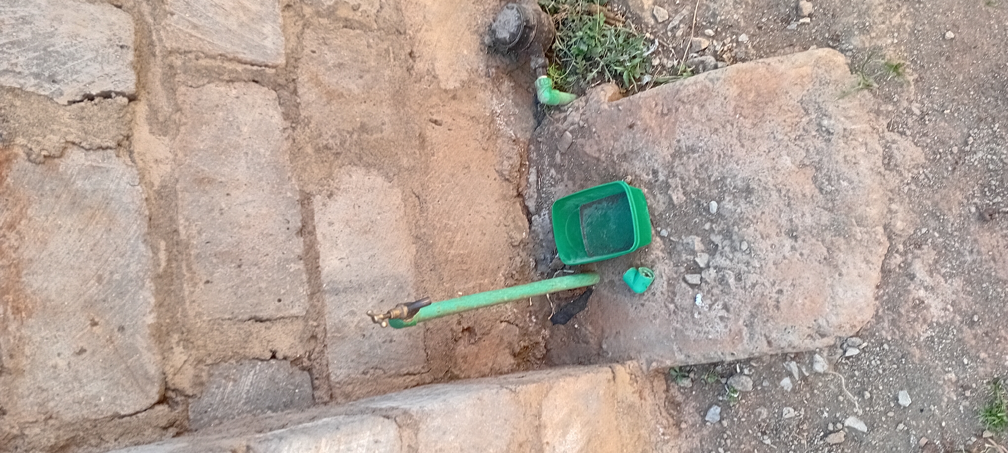 The image depicts an outdoor scene near a brick wall in Kijabe ward, Kenya. In the foreground, a green hose connected to a brass faucet stands vertically. To the right of the faucet, a small, green plastic container is placed on a concrete surface. Nearby, a detached green plastic pipe fitting lies on the ground. The ground is a mixture of dirt, pebbles, and some sparse vegetation. Further to the right, a black circular object, possibly a water meter cover, is visible amidst green grass. The background is dominated by a rough brick wall. No people are present in the image.