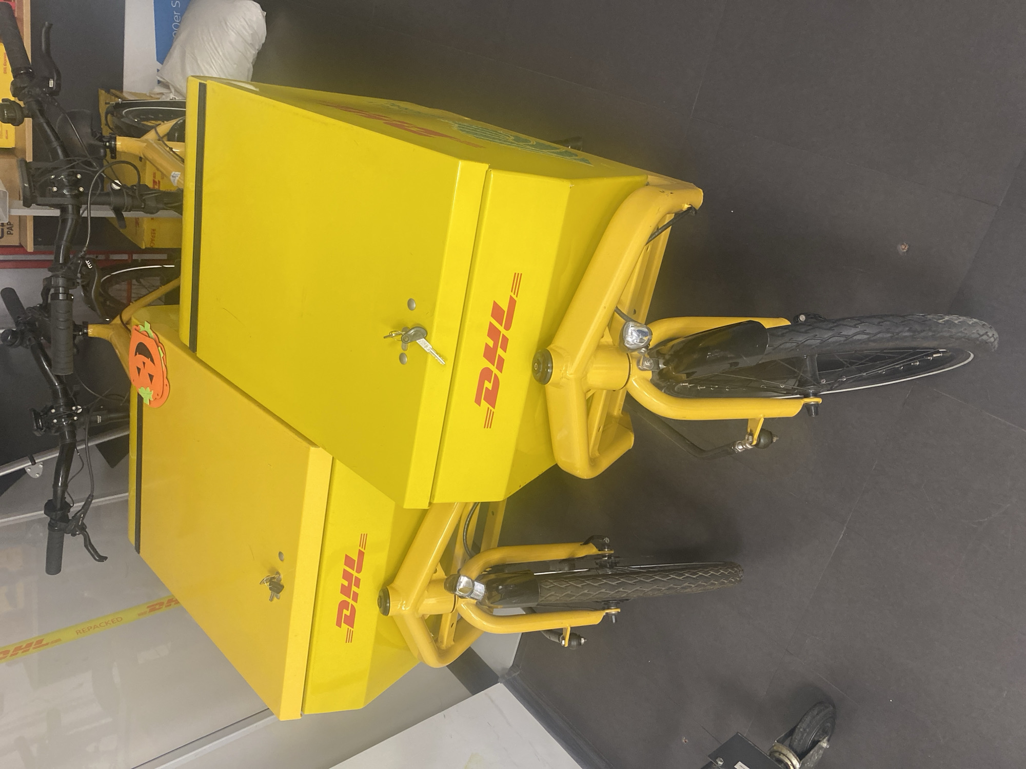A yellow DHL cargo bicycle is the primary subject of the image. The bicycle features two large yellow cargo boxes with the DHL logo prominently displayed. The bicycle appears to be indoors, possibly in a storage area or a loading dock. The background is a combination of white walls and a dark floor. The location cannot be determined to be Sofia, Bulgaria from the visual data alone.